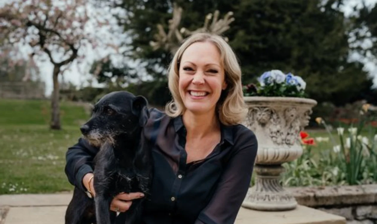 A lady with blonde hair smiles with her arm around a dog