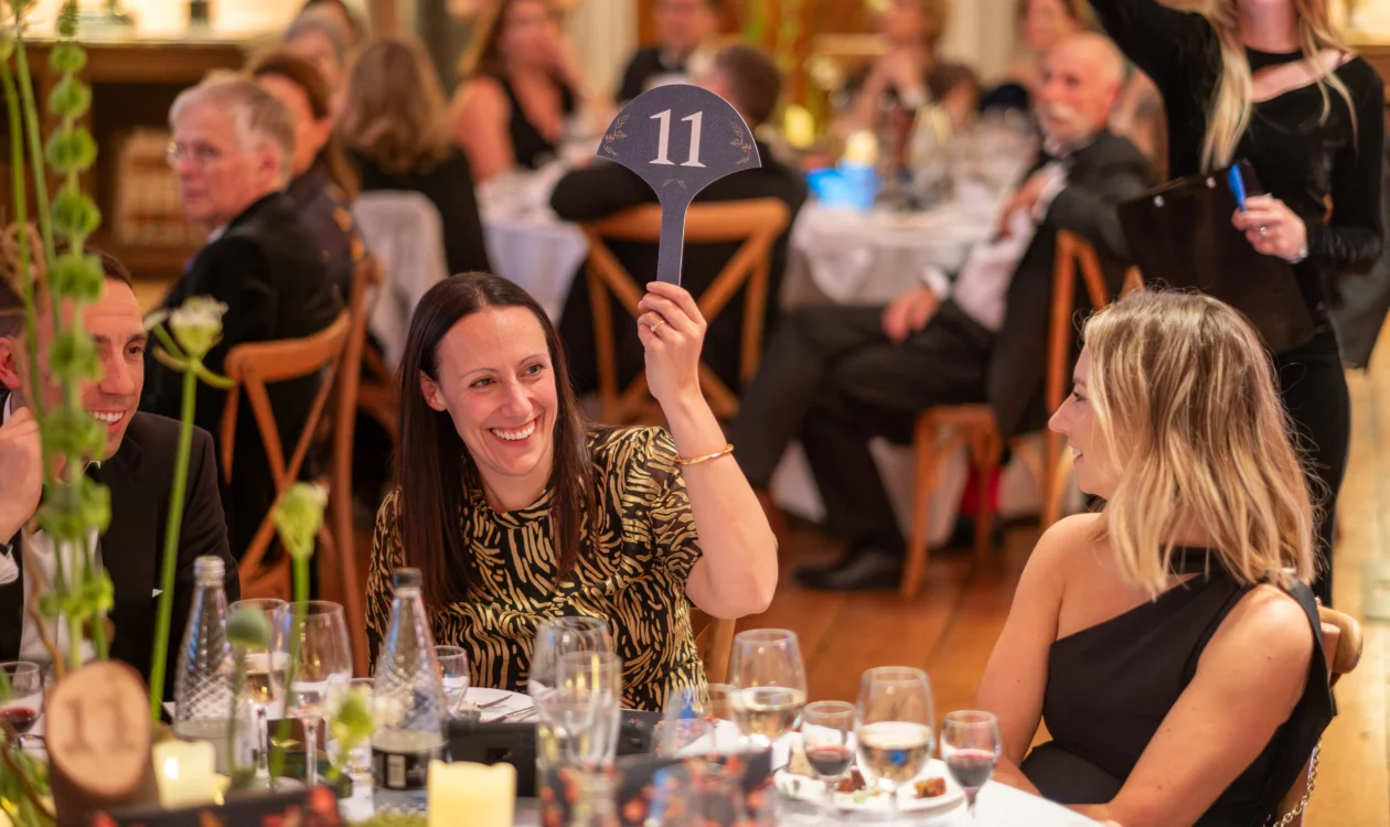 A lady sat at a table at  black tie event raises an auction paddle