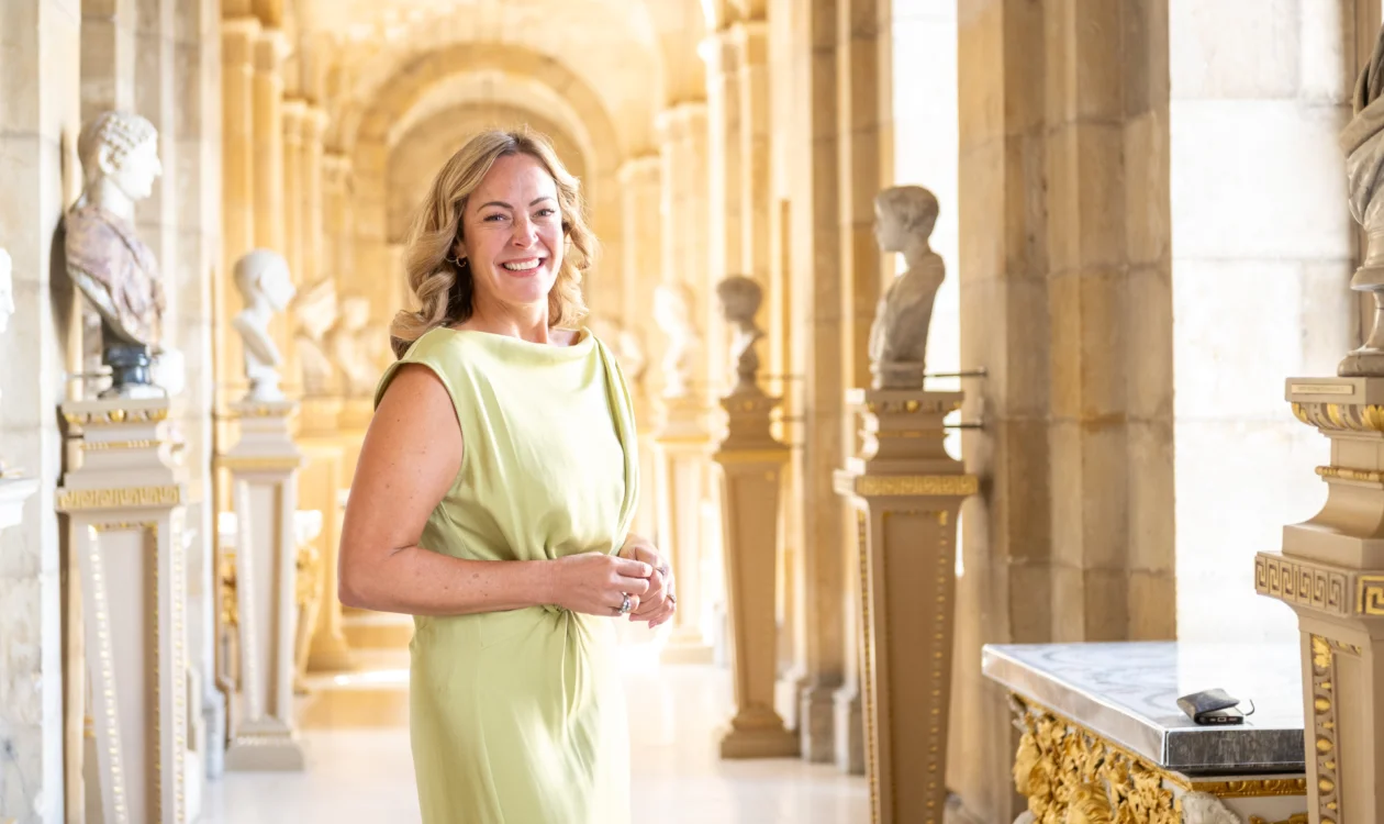 A lady in a pale summer dress smiles in a grand corridor