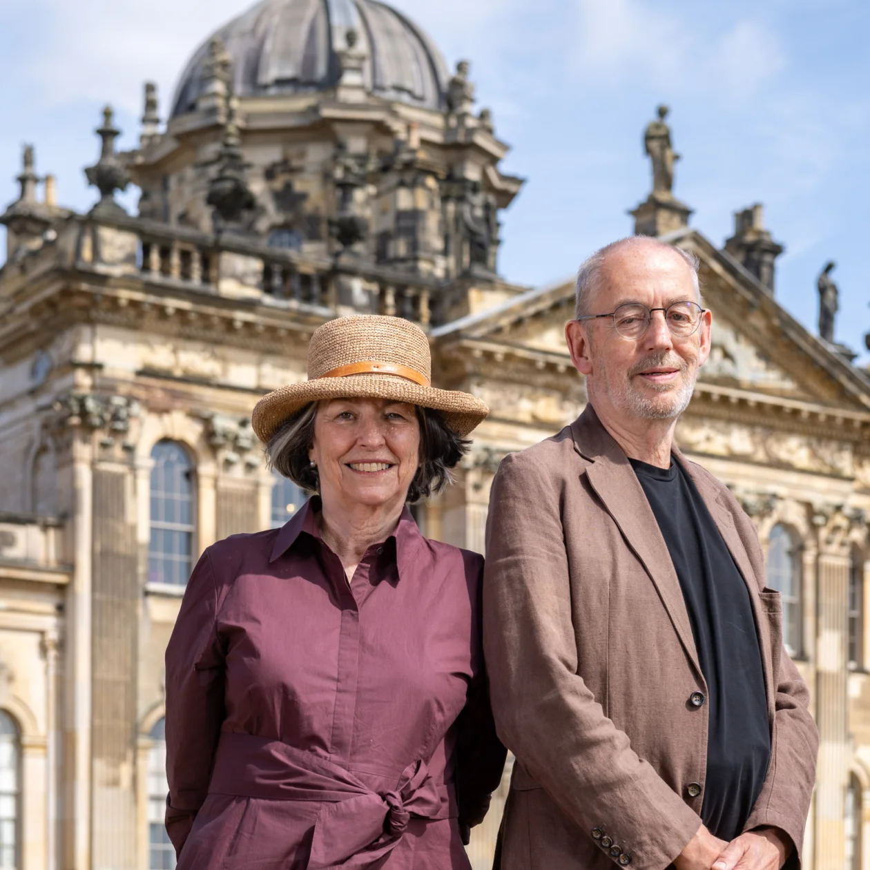 A lady in a sun hat and a man in a brown linen suit stand in front of a stately home