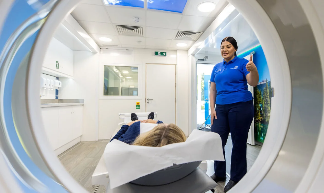 Looking through a CT scanner to see a nurse talking to a patient who is lying down, ready for their scan