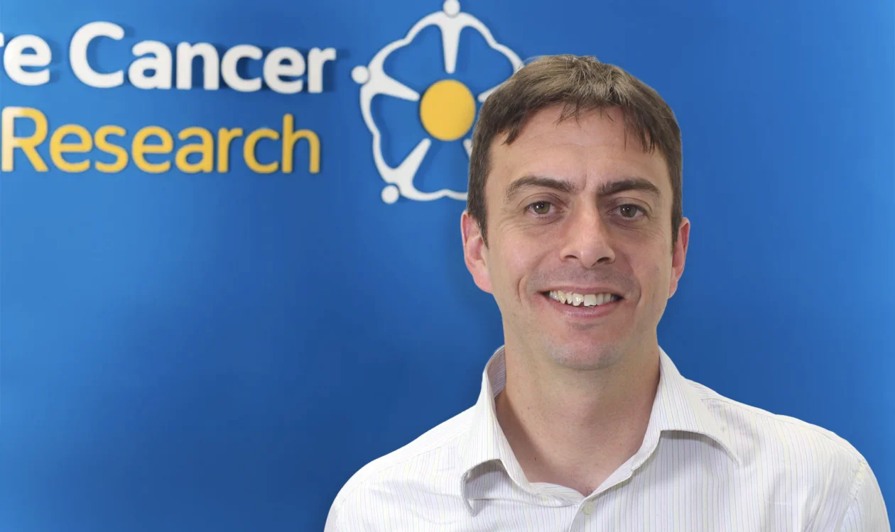 A man stands in front of a Yorkshire Cancer Research logo on a wall