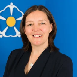 Kathryn Scott standing in front of a blue backdrop smiling at the camera. Behind her is the charity's logo.