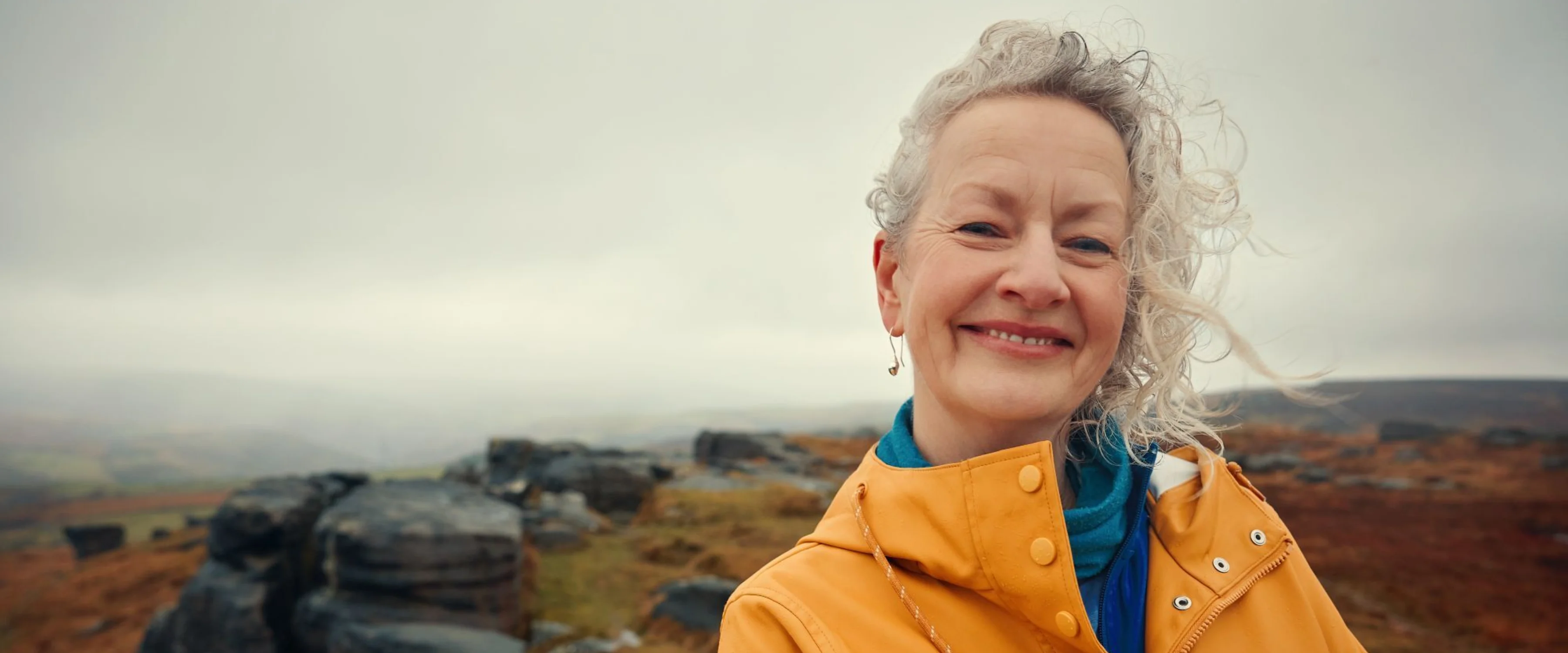 A lady in a yellow waterproof stands in the wind on a Yorkshire hilltop