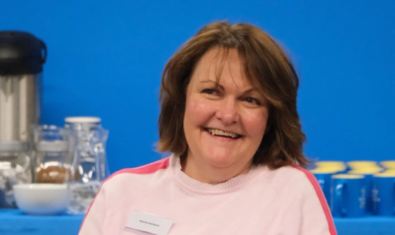 A woman in a pink jumper smiles across a room