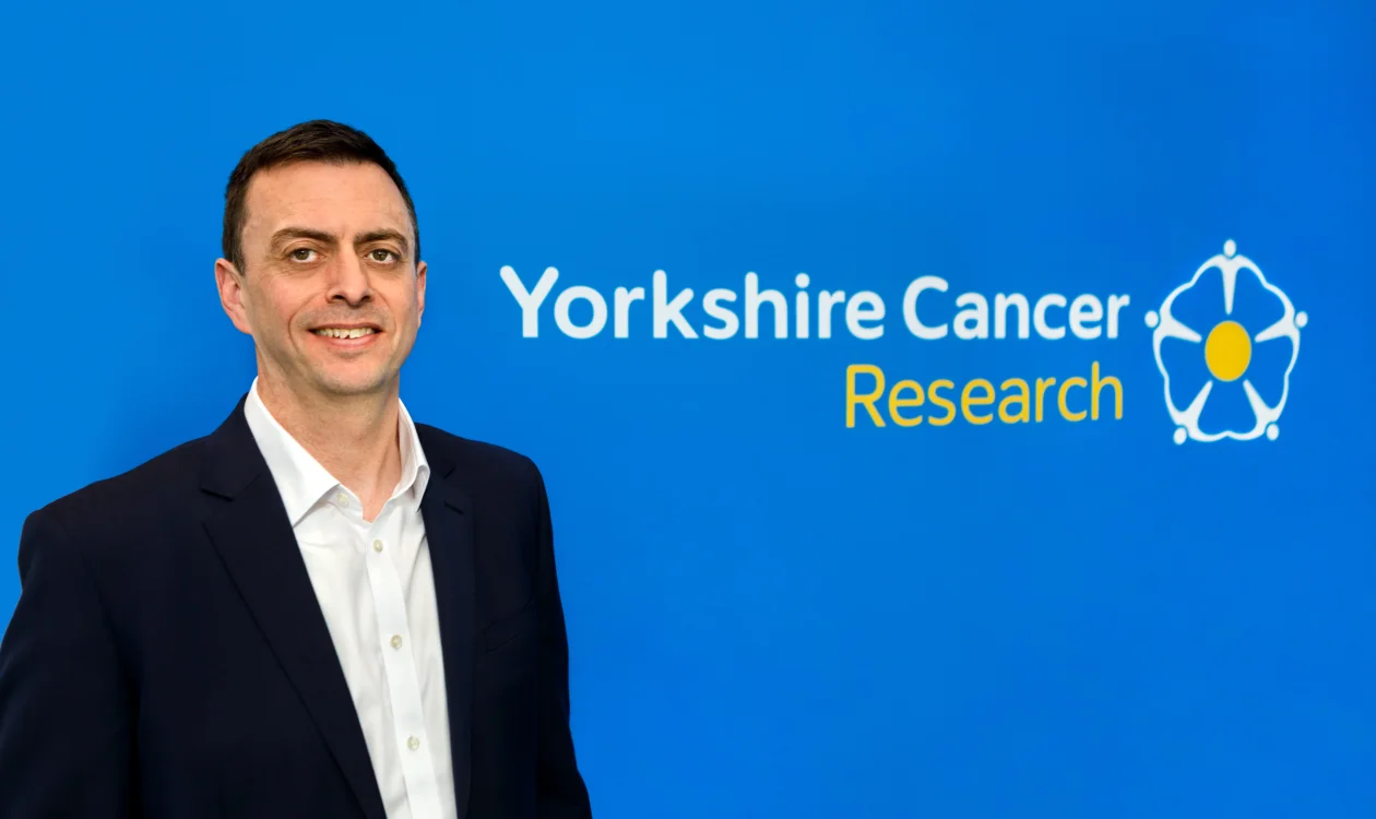 A man in a blazer stands in front of a Yorkshire Cancer Research logo