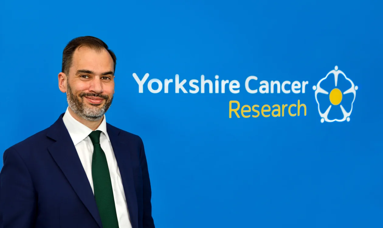 A man in suit and tie stands in front of a Yorkshire Cancer Research logo