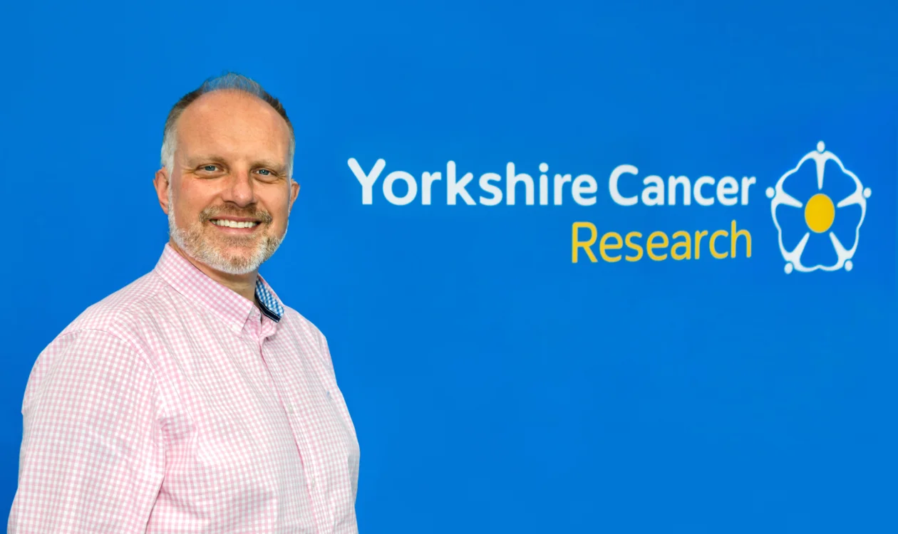 A man in a pink shirt stands in front of a Yorkshire Cancer Research logo