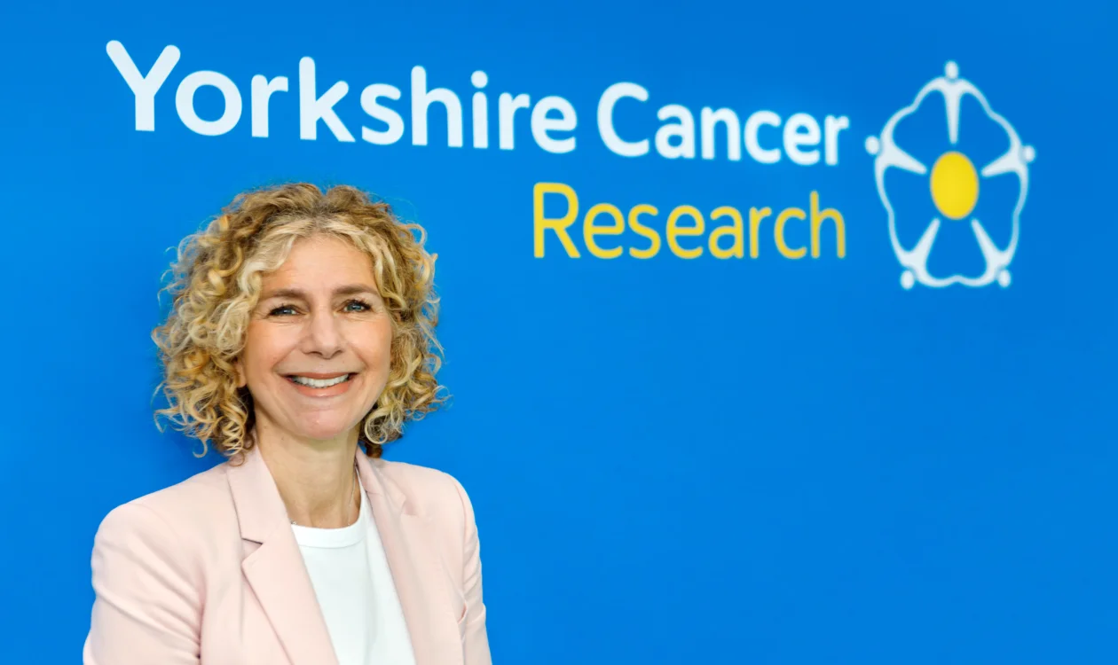A lady in a pink blazer stands in front of a Yorkshire Cancer Research logo