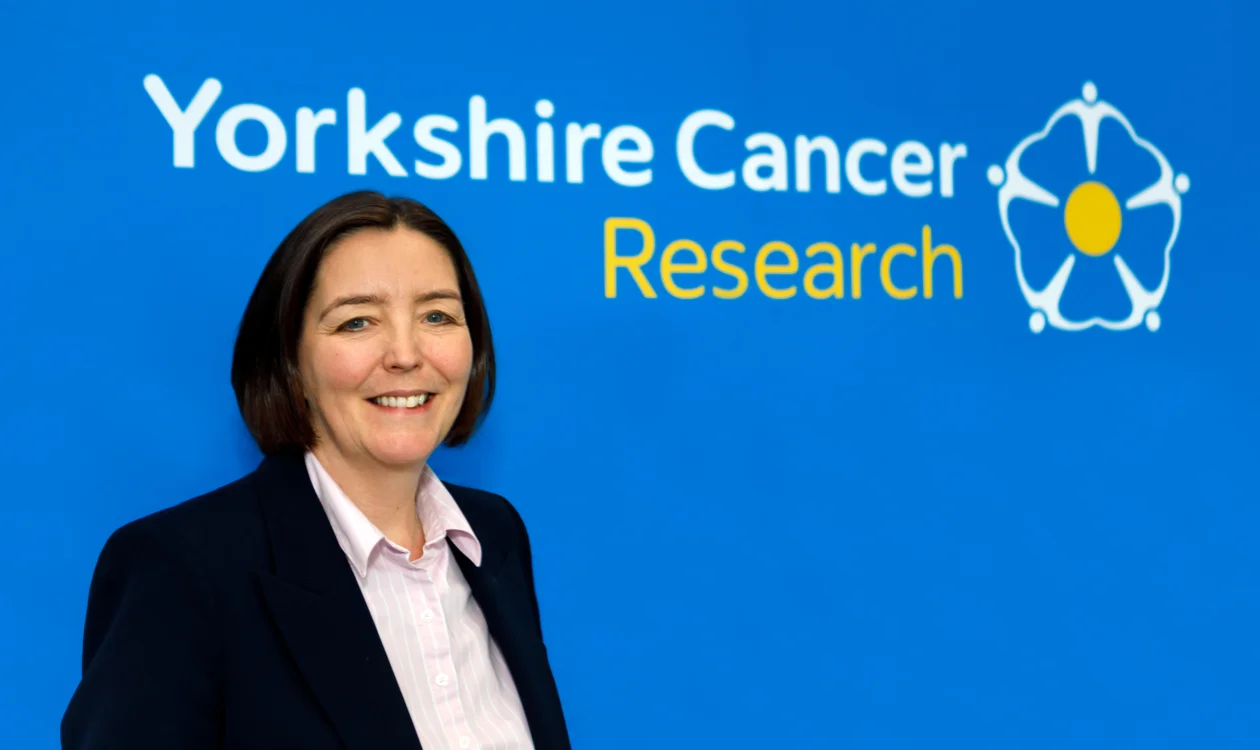 A lady in a dark blazer stands in front of a Yorkshire Cancer Research logo