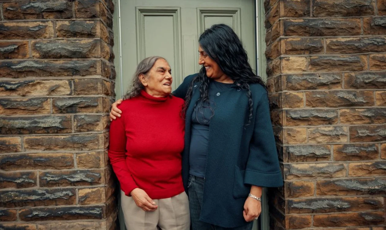A mum and daughter stand at a front door smiling at each other. The daughter has her arm around her mum.