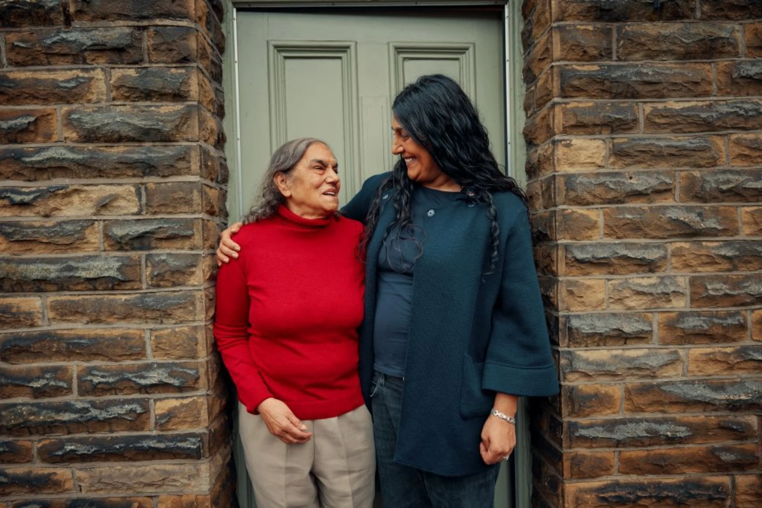 A mum and daughter stand at a front door smiling at each other. The daughter has her arm around her mum.