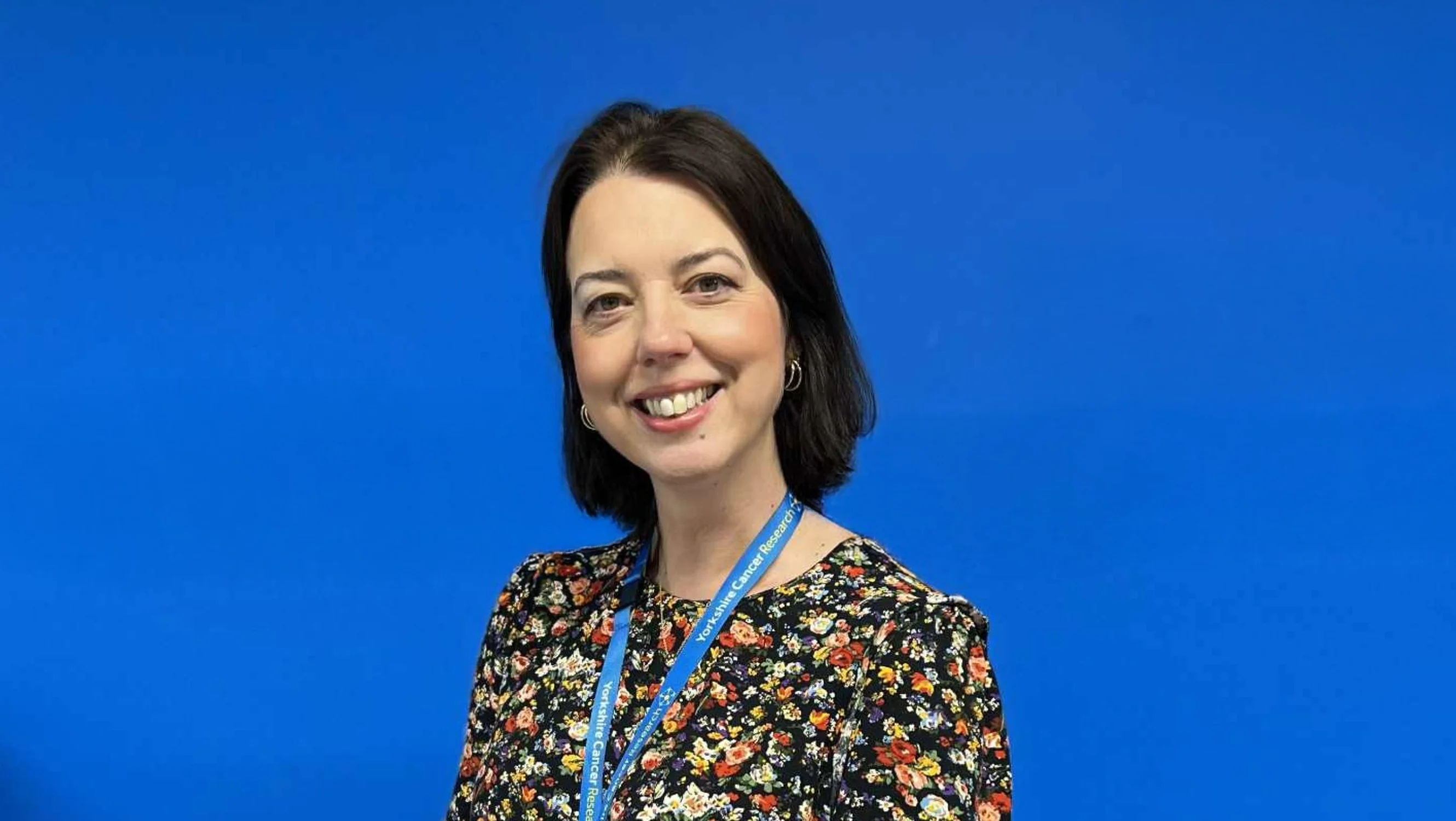 A lady in floral blouse smiles in front of a blue wall