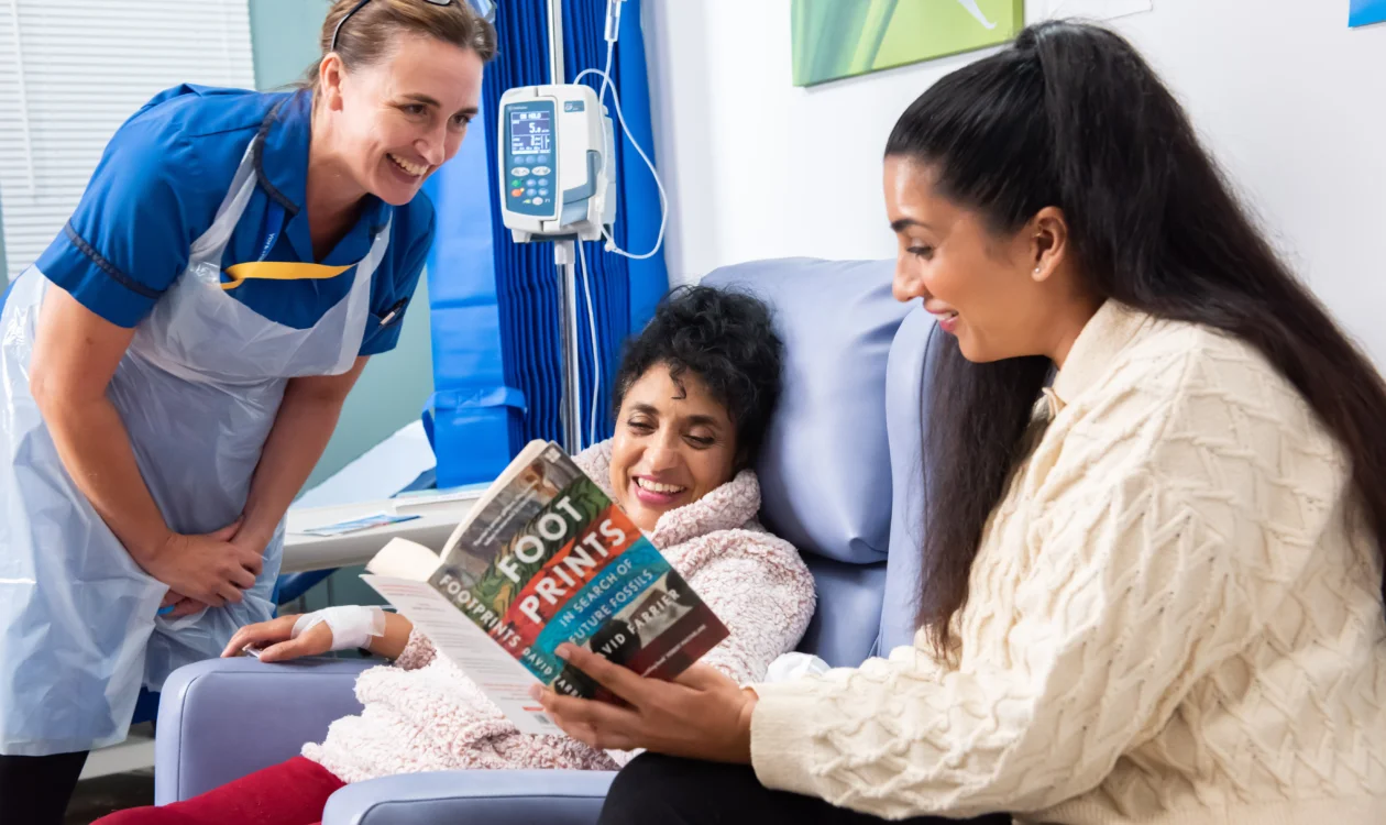 Female cancer patient on a hospital ward with daughter visiting her and reading to her. A nurse is standing next to them.
