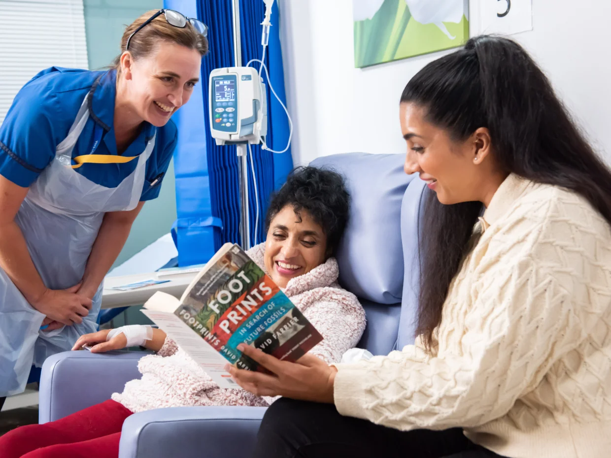 Female cancer patient on a hospital ward with daughter visiting her and reading to her. A nurse is standing next to them.