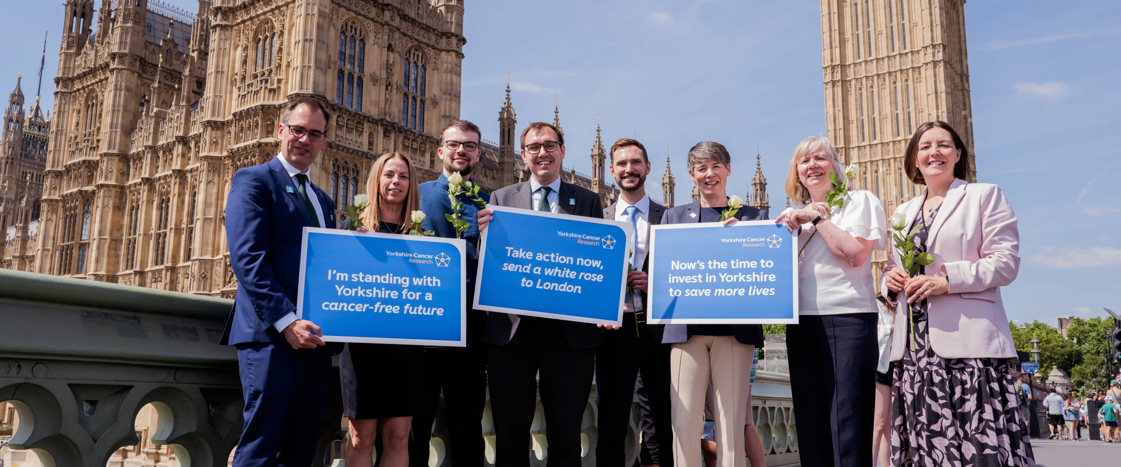 A group of people stand in front of Westminster holding white roses and placards of support for Yorkshire Cancer Research