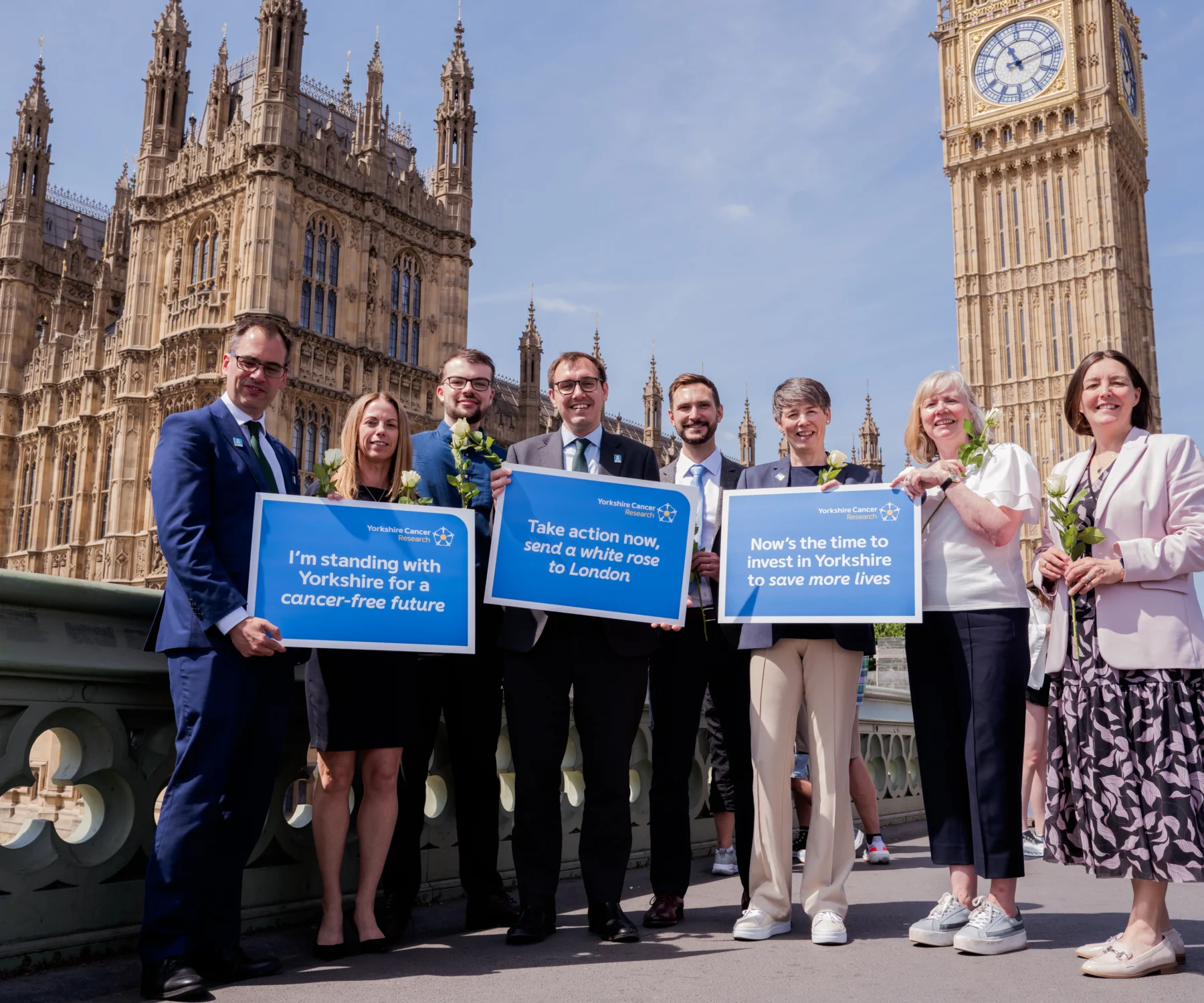 A group of people stand in front of Westminster holding white roses and placards of support for Yorkshire Cancer Research