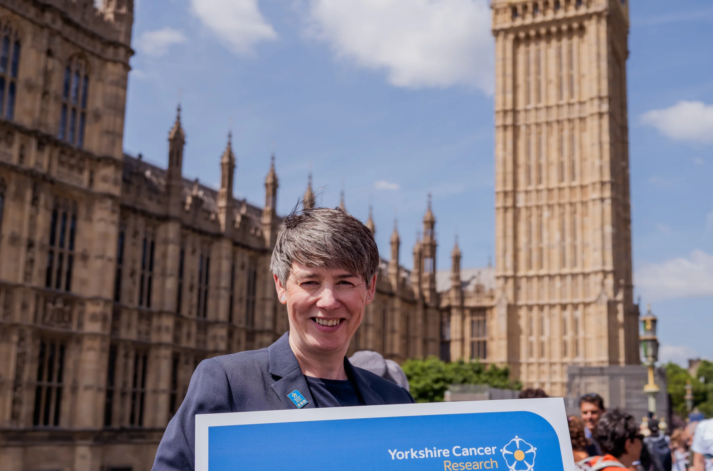 A Yorkshire Cancer Research supporter holding a placard and a rose in front of Houses of Parliament. The Placard reads 'Now's the time to invest in Yorkshire to save more lives'.