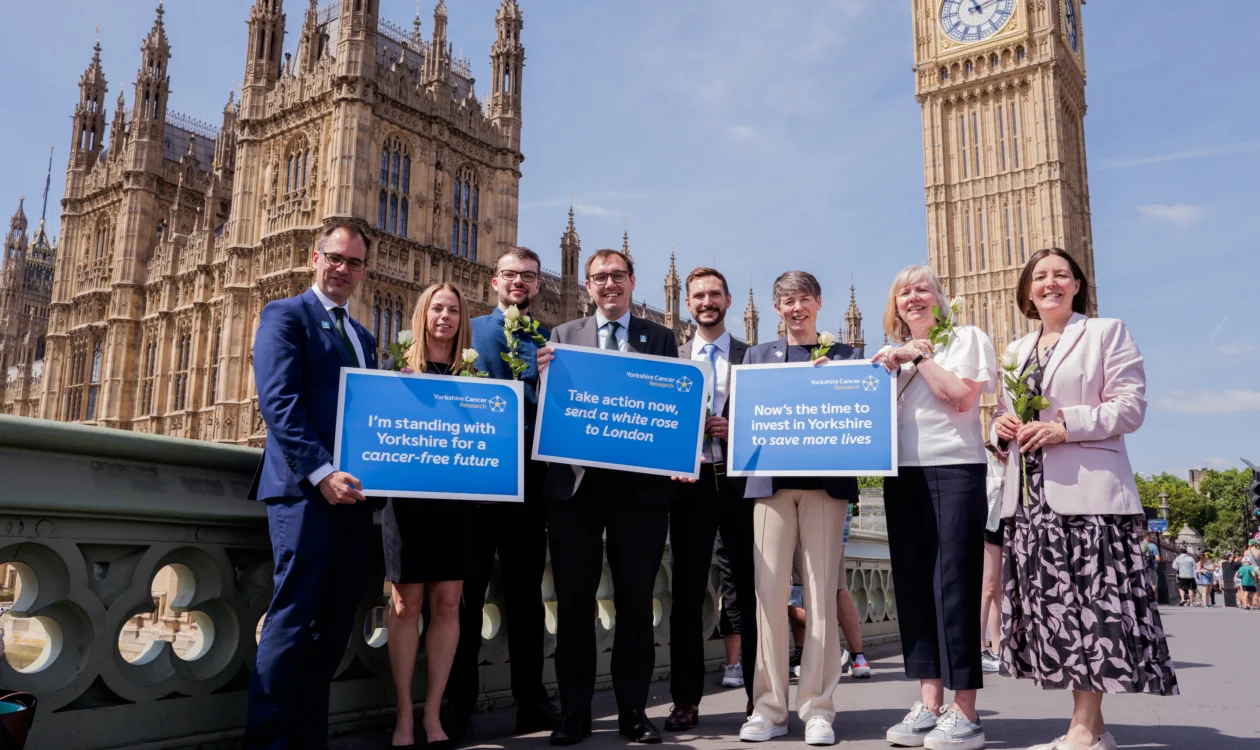 A group of people stand in front of Westminster holding white roses and placards of support for Yorkshire Cancer Research