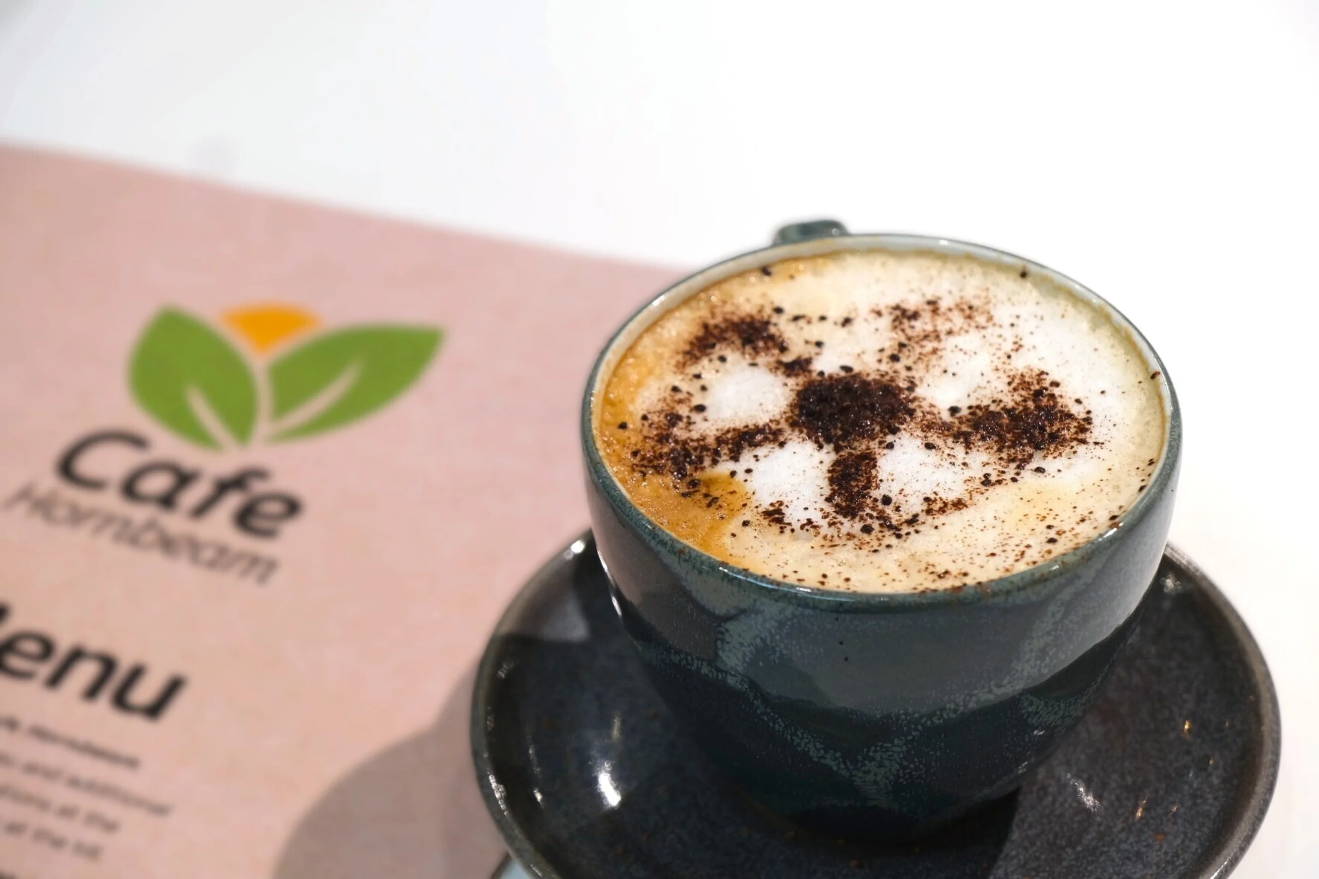 A frothy cappuccino in the cafe with a dusting of chocolate on the top in the shape of the Yorkshire Cancer Research logo. Behind it is a copy of the physical cafe menu.