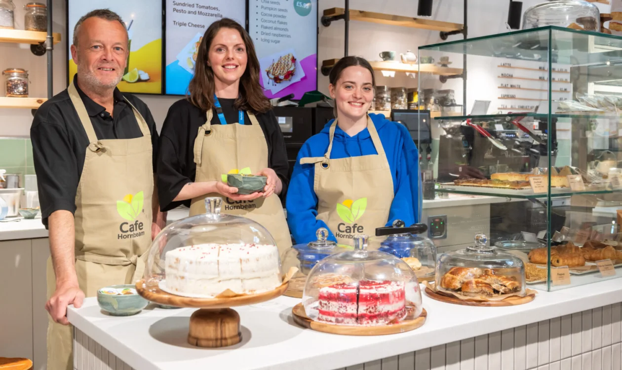 A man and two women in aprons stand at a counter in front of cakes