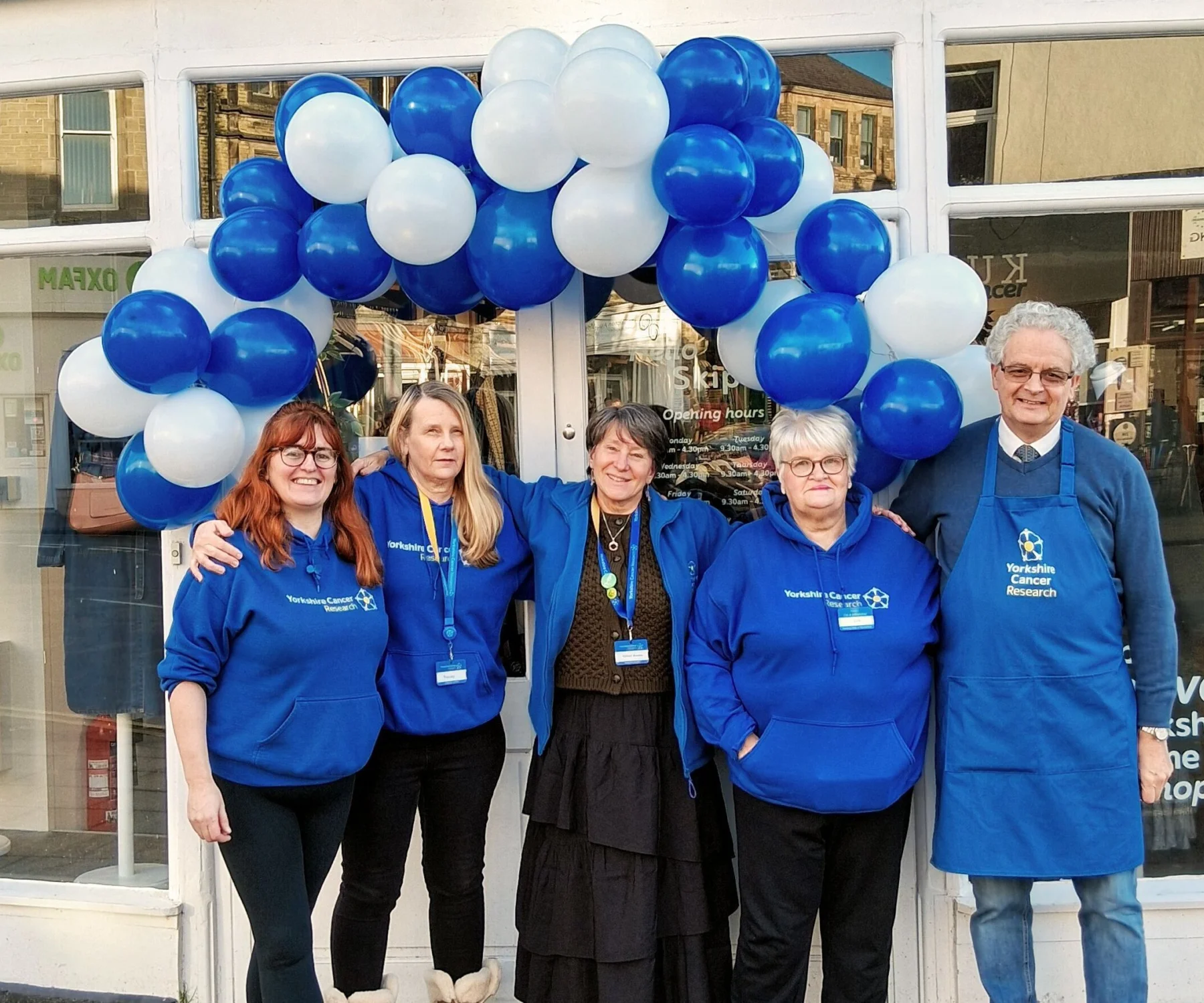 Five people wearing blue hoodies and aprons standing outside the entrance of the Skipton Keighley Road shop. Above them is a blue and white balloon arch.