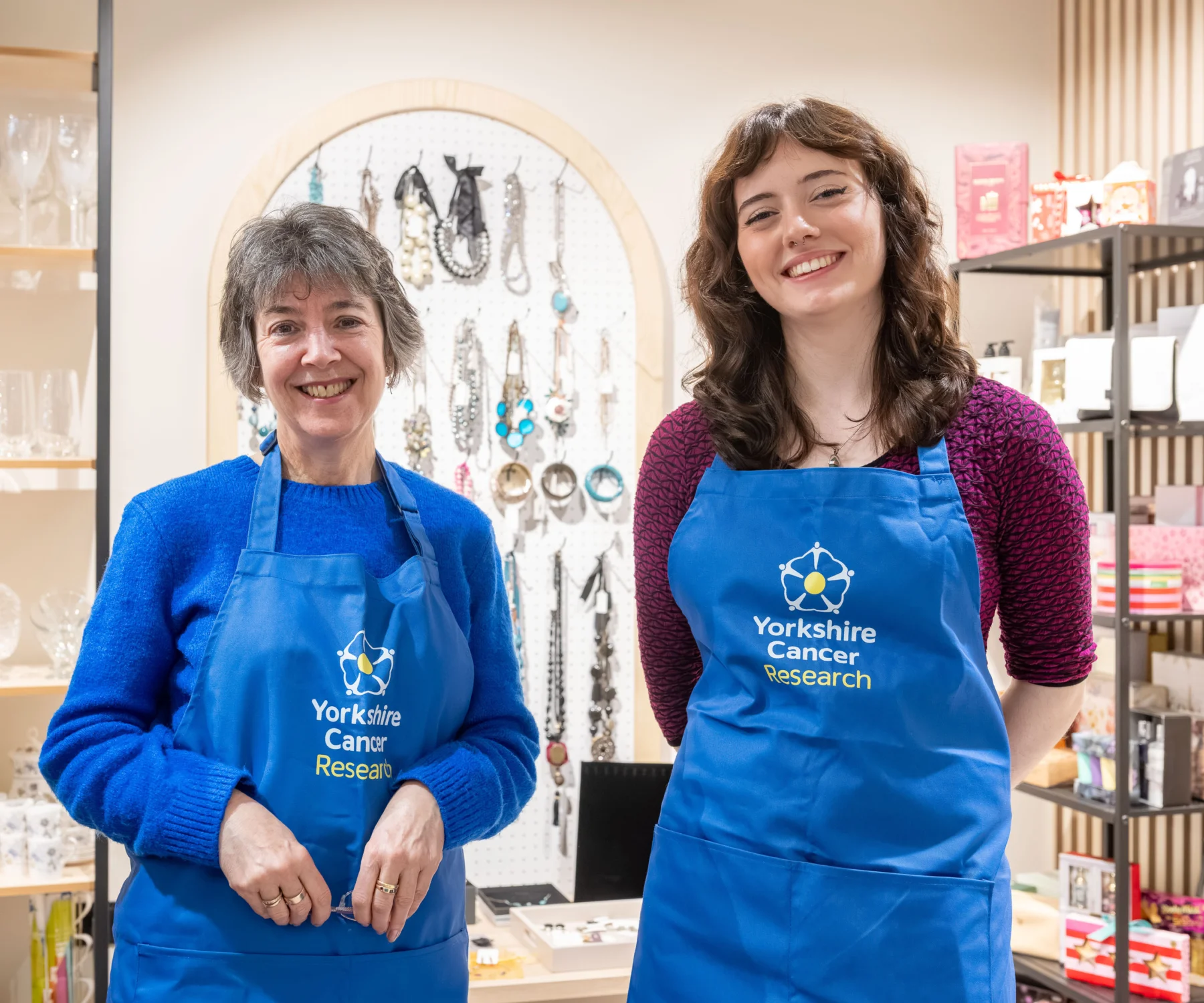 Two ladies wearing blue "Yorkshire Cancer Research" aprons are stood in a retail setting. Behind them is a pinboard display with jewellery on it.