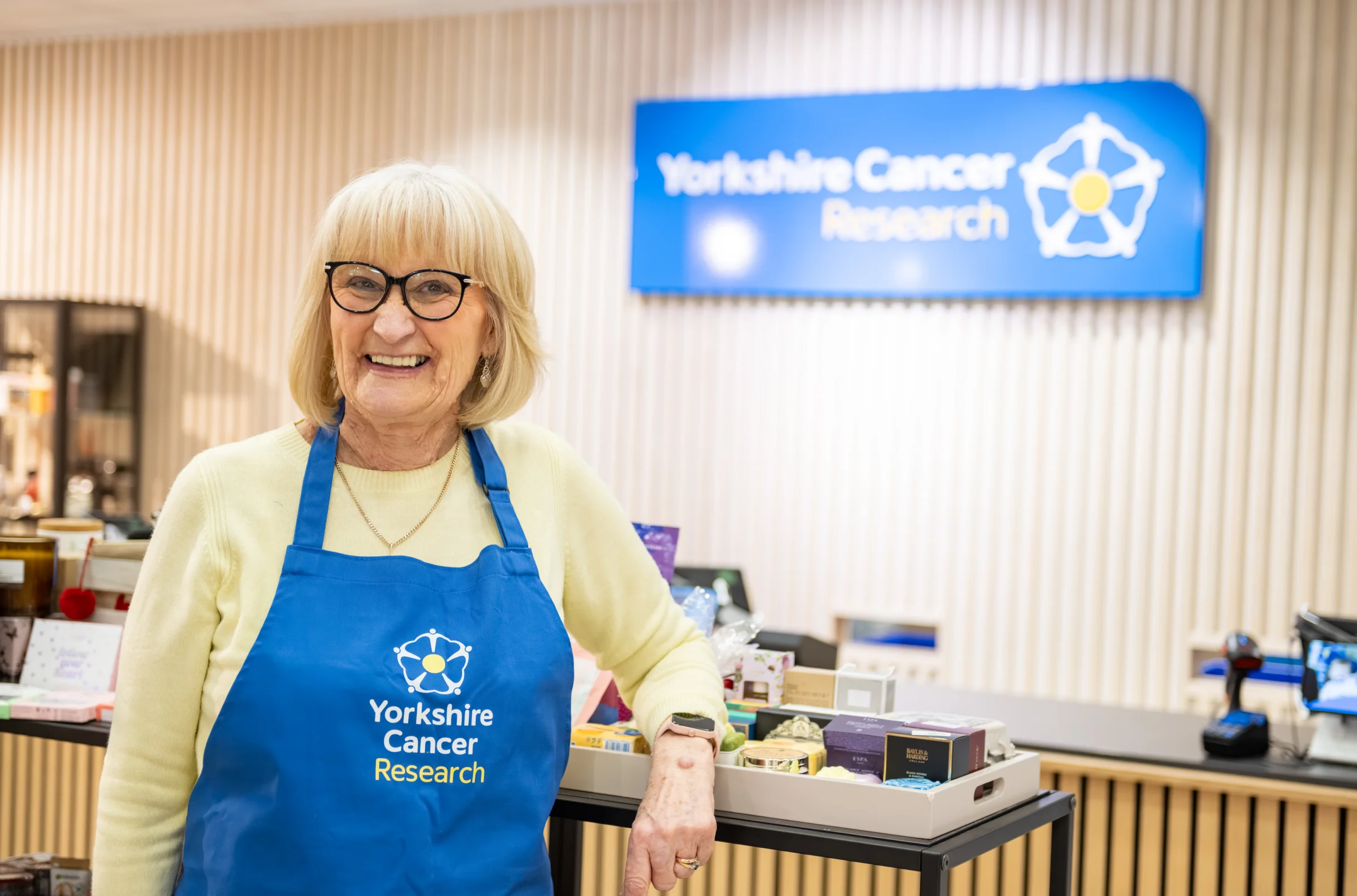 A lady wearing black glasses, a yellow jumper and a blue Yorkshire Cancer Research apron is leaning on a shop display. Behind her is a sign with the charity's logo and a till area.