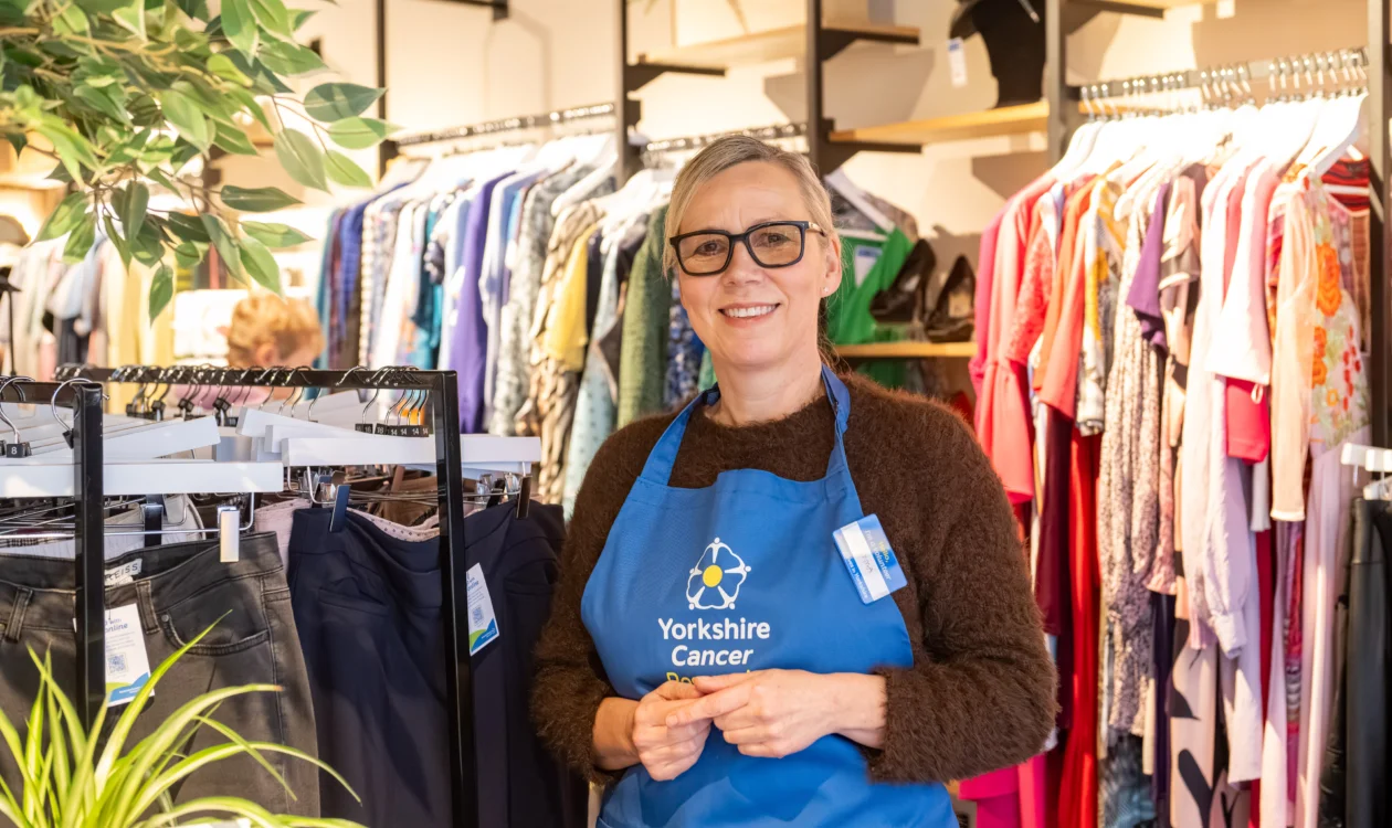 A female retail volunteer wearing glasses and a blue "Yorkshire Cancer Research" apron is stood in a shop. Behind her are rails of ladies clothing and fake plants on shelves.