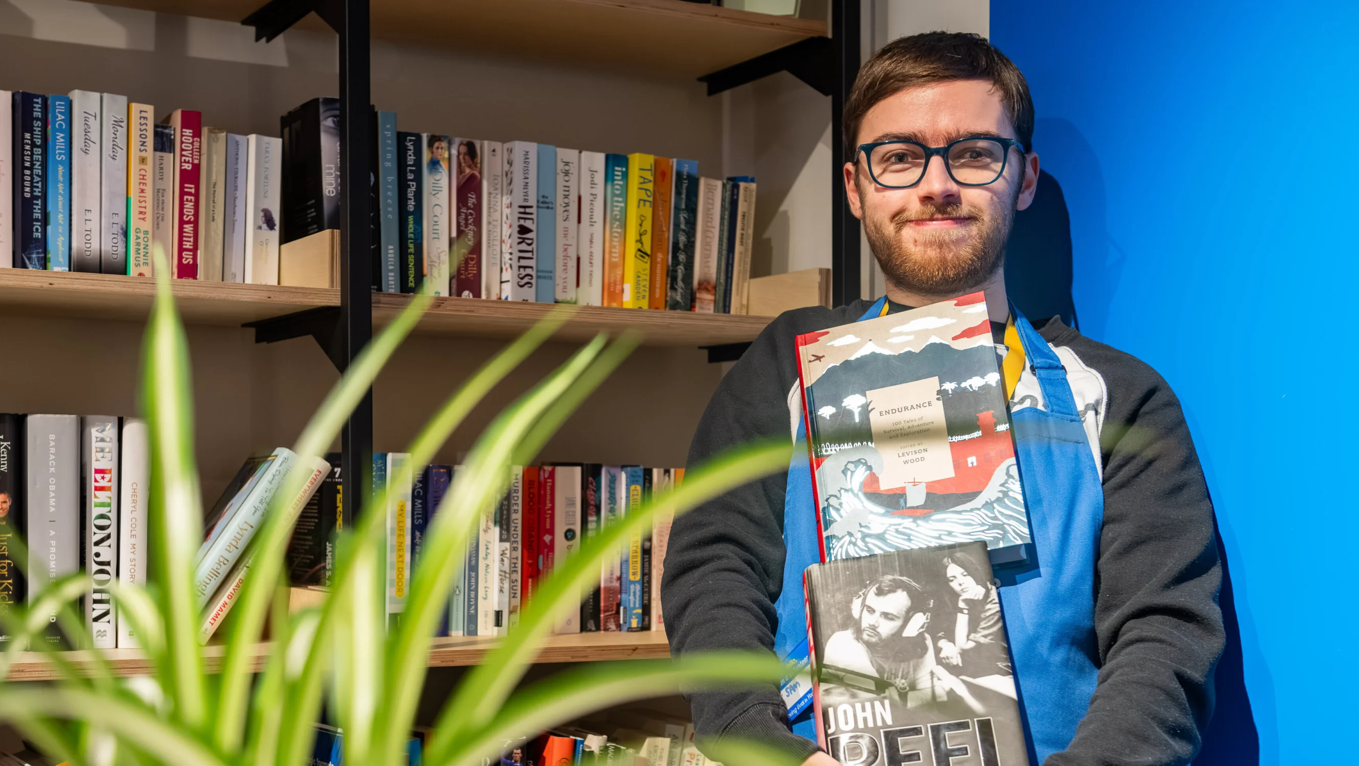A young man wearing a blue apron is stood holding a pile of fiction books. Behind his right shoulder is a blue wall, and on the other side behind him is a bookshelf in a retail setting. In the foreground is a blurred indoor plant.