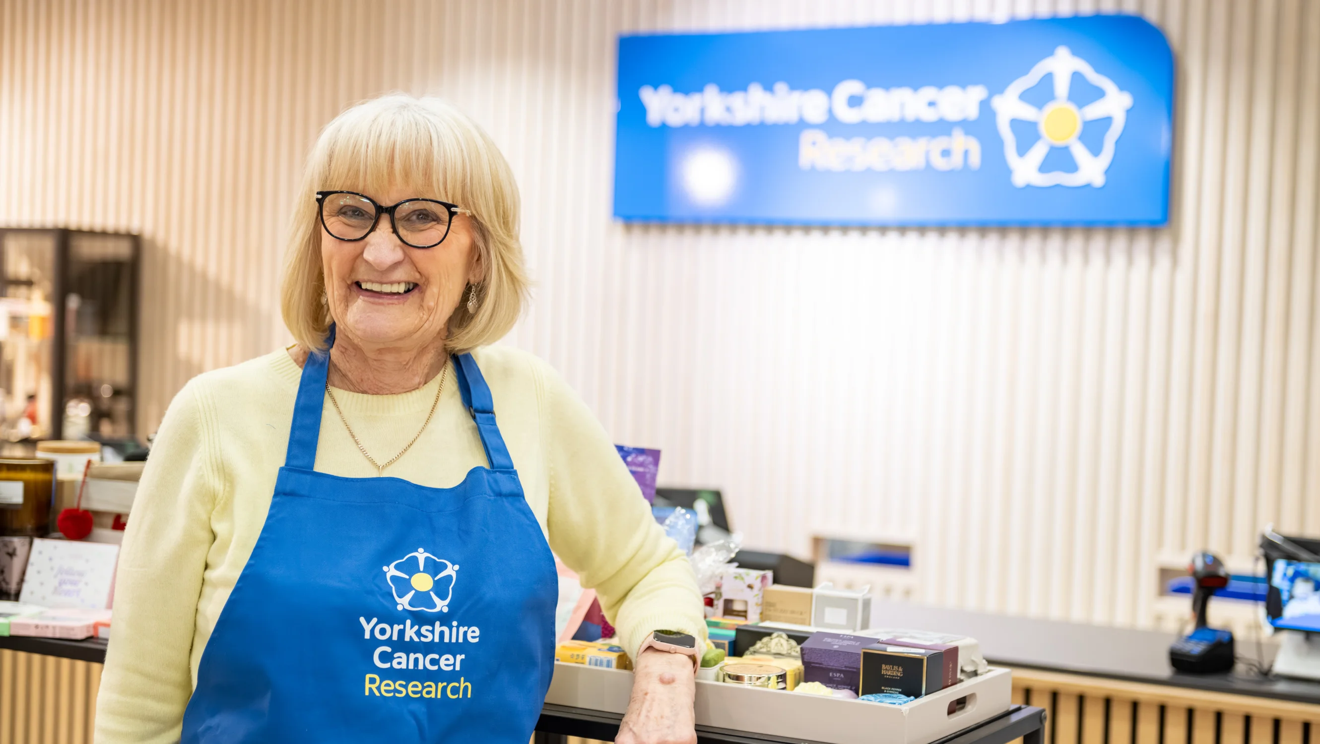 A lady wearing black glasses, a yellow jumper and a blue Yorkshire Cancer Research apron is leaning on a shop display. Behind her is a sign with the charity's logo and a till area.