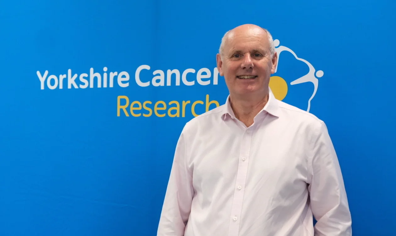 A man in a shirt stands in front of the Yorkshire Cancer Research logo