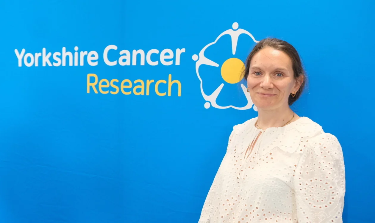 A lady in a white top stands in front of a Yorkshire Cancer Research logo