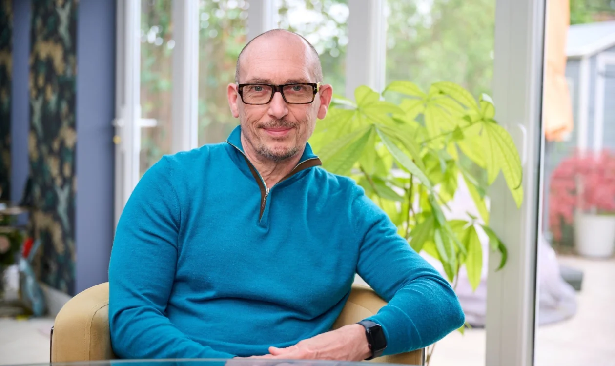 A man in a blue ip up top and glasses smiles on a chair