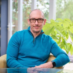 A man with glasses sits in a chair smiling