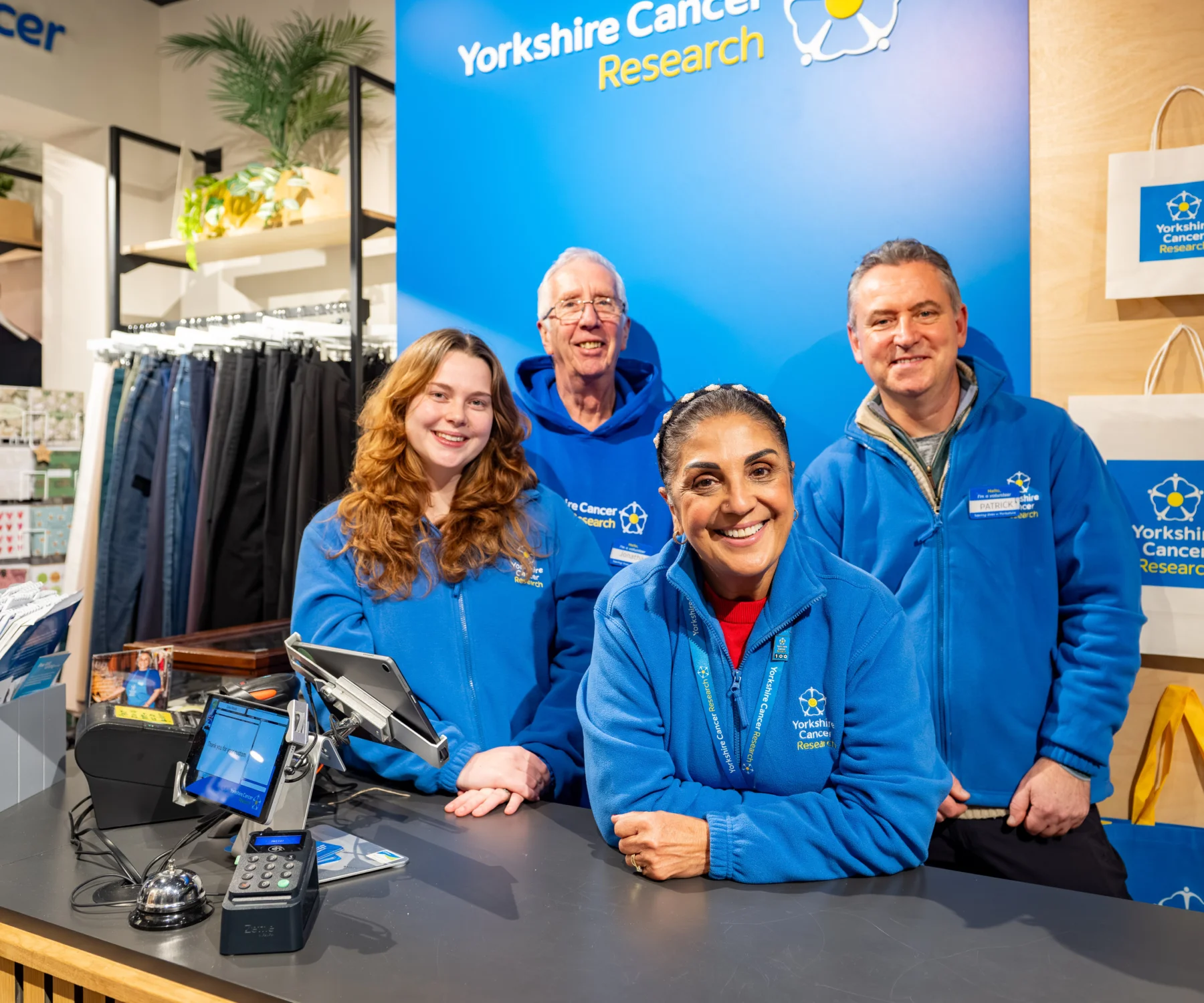Four people are stood behind a till at a Yorkshire Cancer Research shop. Behind them is a blue wall and they are all wearing branded blue jumpers with the charity's logo on.