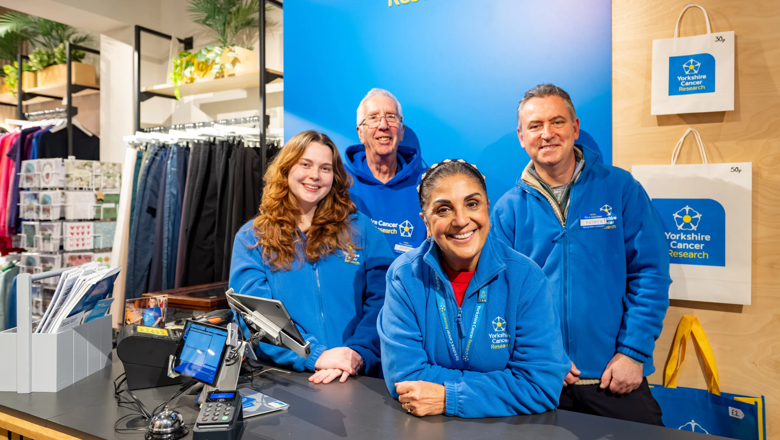 Four people are stood behind a till at a Yorkshire Cancer Research shop. Behind them is a blue wall and they are all wearing branded blue jumpers with the charity's logo on.