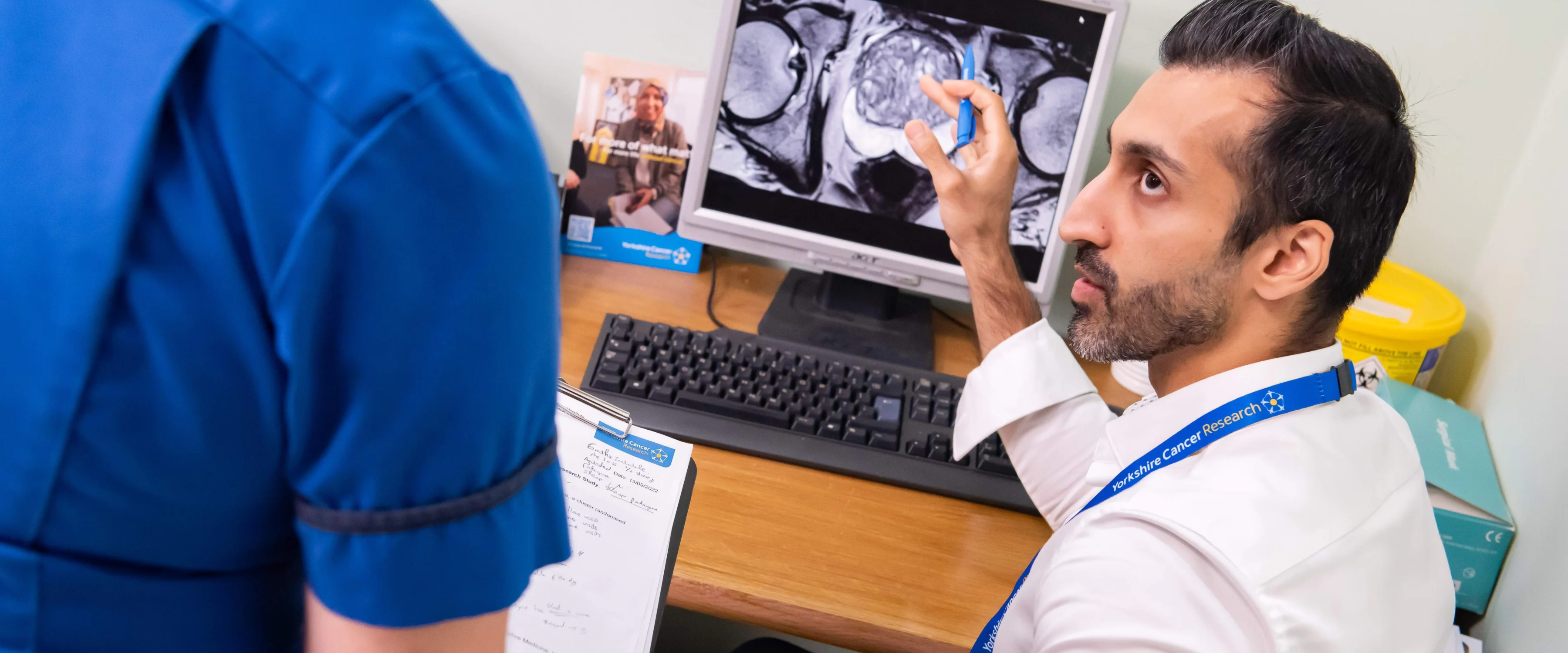 A medical professional sat at a desk speaks to a nurse next to him about a scan on a computer screen