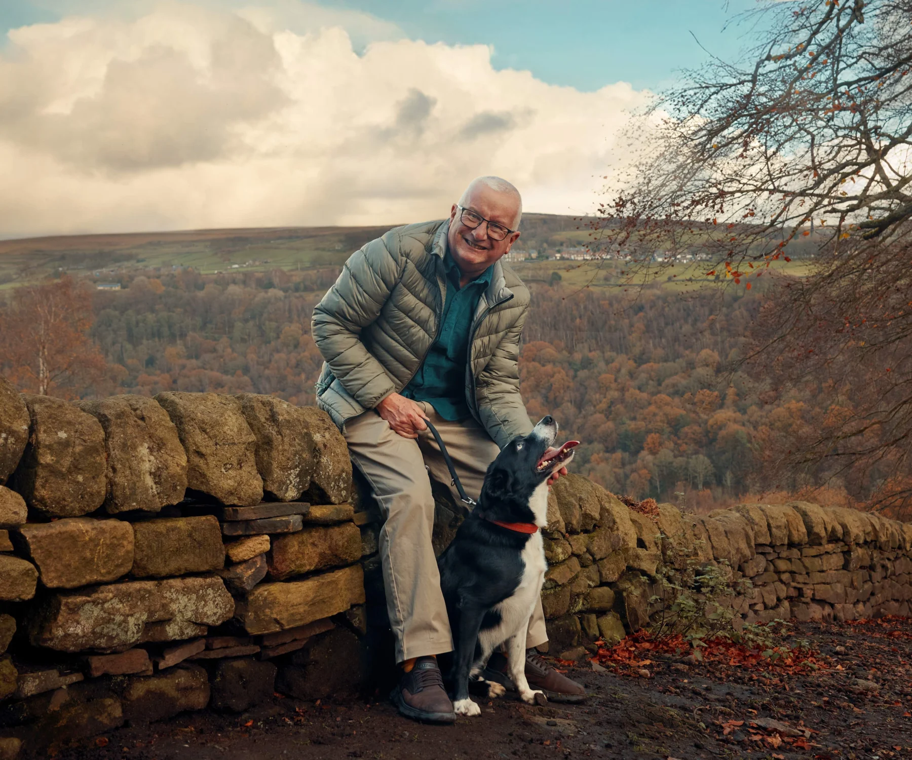 A man sits on a stone wall smiling at the camera, with a border collie dog at his feet