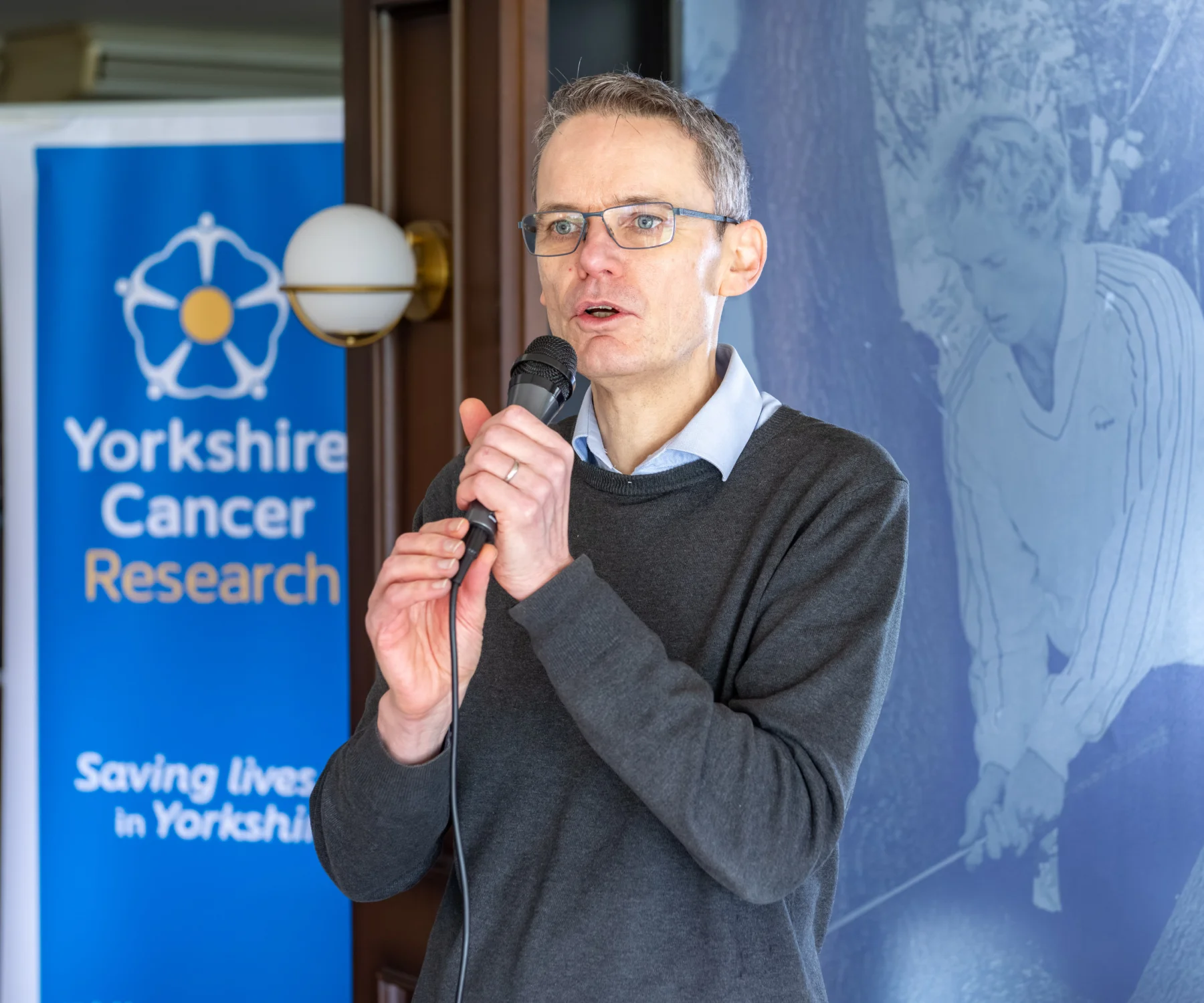 Andy Wilson is giving a talk in front of a Yorkshire Cancer Research banner.