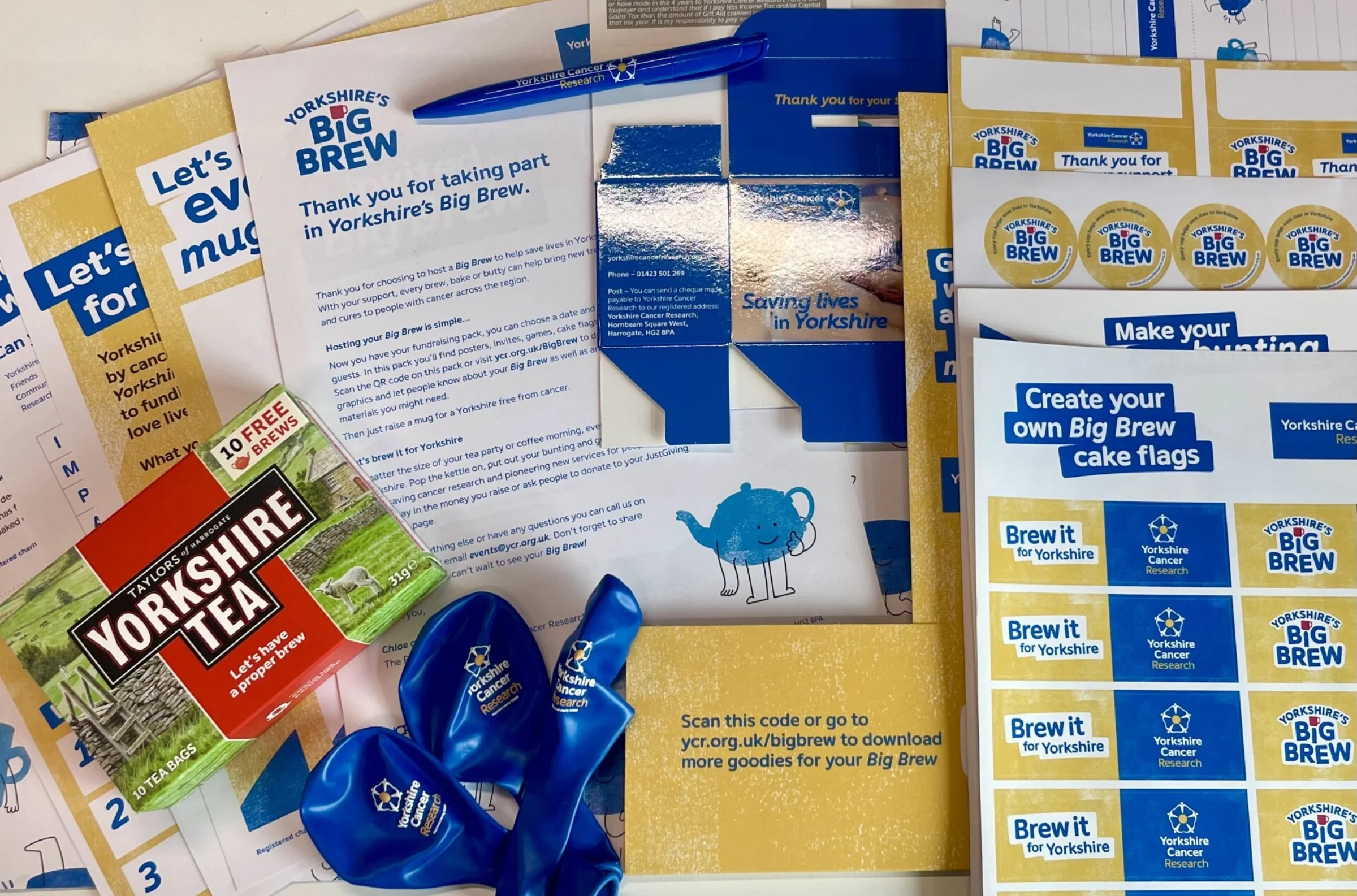The Big Brew fundraising pack contents are laid out. There's various sheets of paper including "Create your own cake flags" and a sticker sheet. There's a box of Yorkshire tea, blue pen,  three blue balloons and a cardboard collection box also visible.