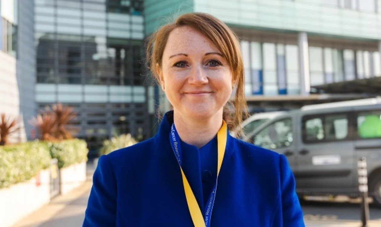 Jenny is wearing a blue coat and a yellow lanyard smiling at the camera outside a modern building.