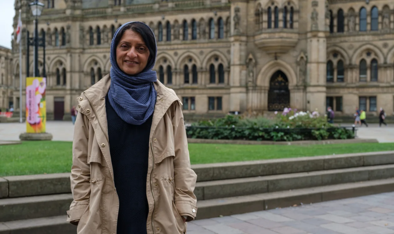 Dr Aliya Darr is wearing a blue headscarf and a beige trench coat, standing in front of the Bradford Town Hall.