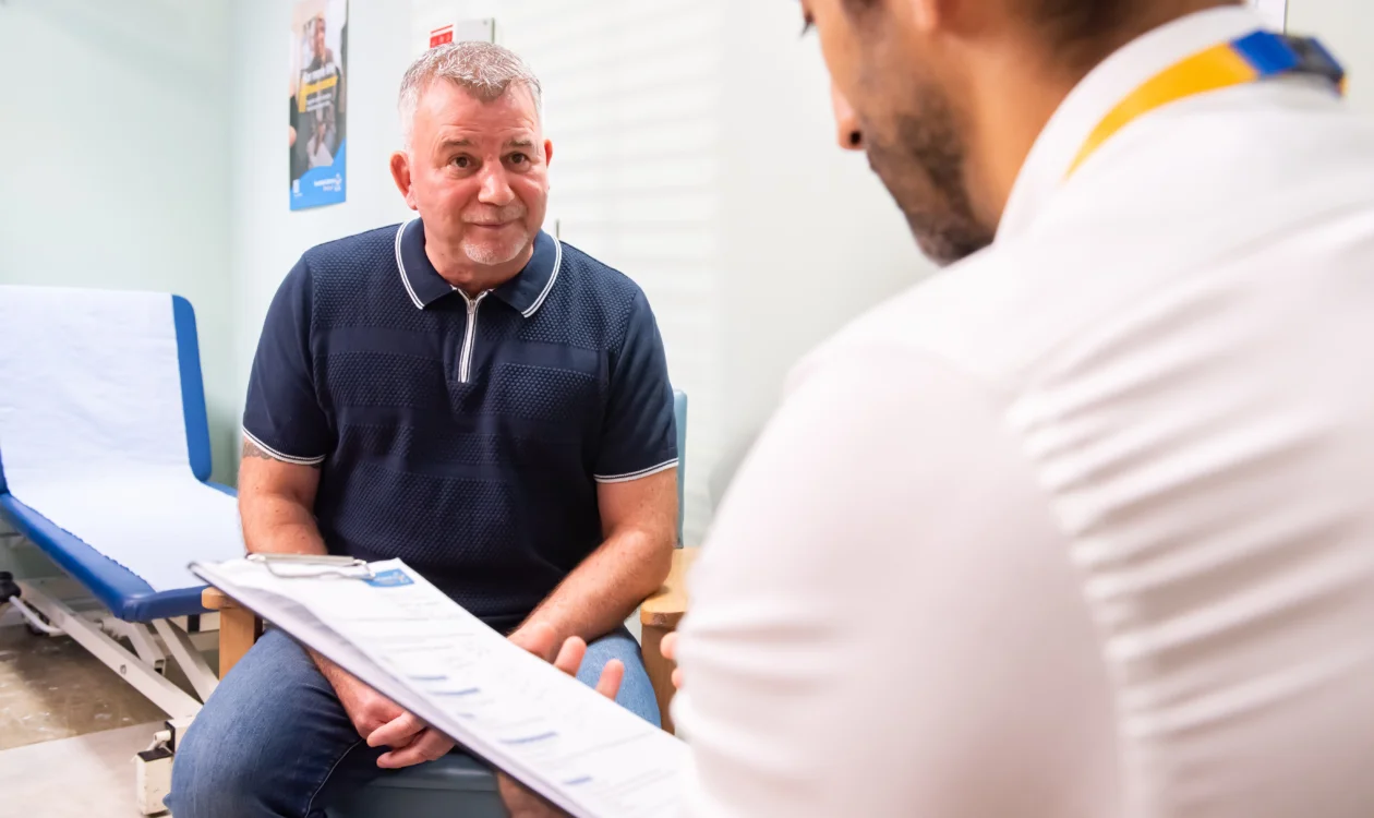 A patient sits in a medical setting while in conversation with a healthcare professional, who is wearing a Yorkshire Cancer Research lanyard and holding a clipboard.
