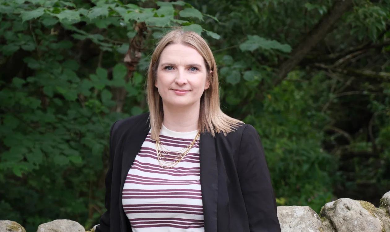 Beth Elias standing, facing the camera in a striped shirt, black blazer against a wall and trees