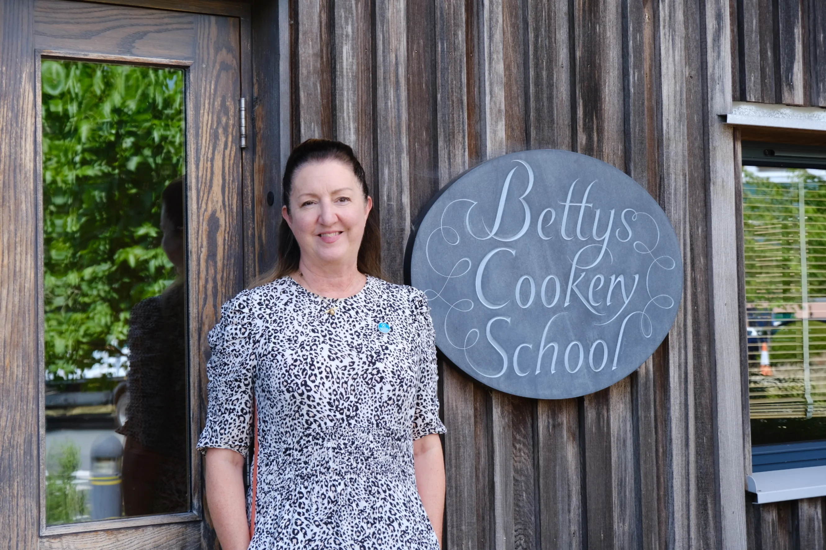Ruth stands outside Bettys Cookery School in Harrogate, wearing a patterned dress and smiling at the camera
