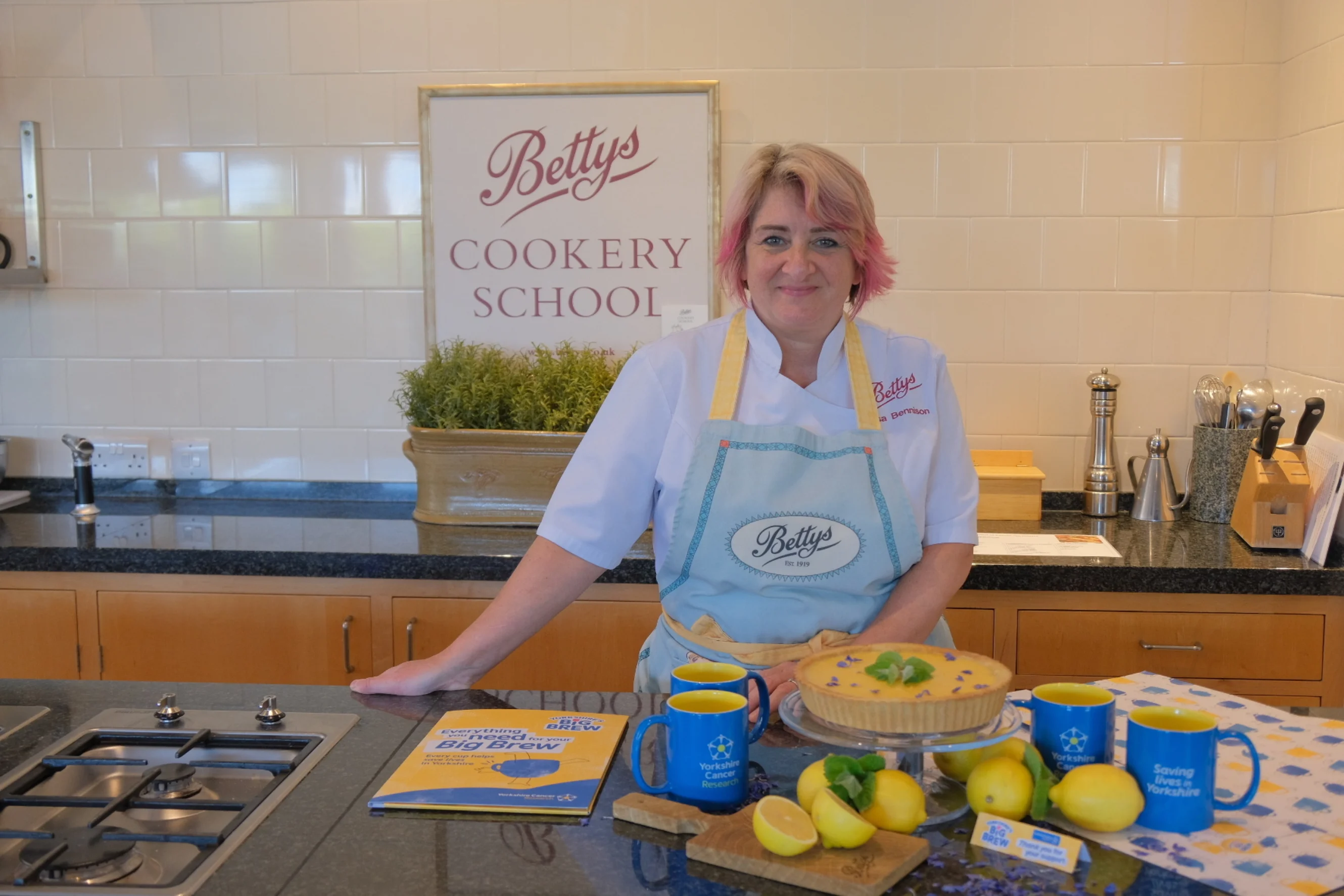 Lisa stands inside Bettys Cookery School, wearing a branded apron and smiling at the camera. On the worktop in front of her is a lemon tart, surrounded by lemons, Yorkshire Cancer Research mugs and a Big Brew fundraising pack.