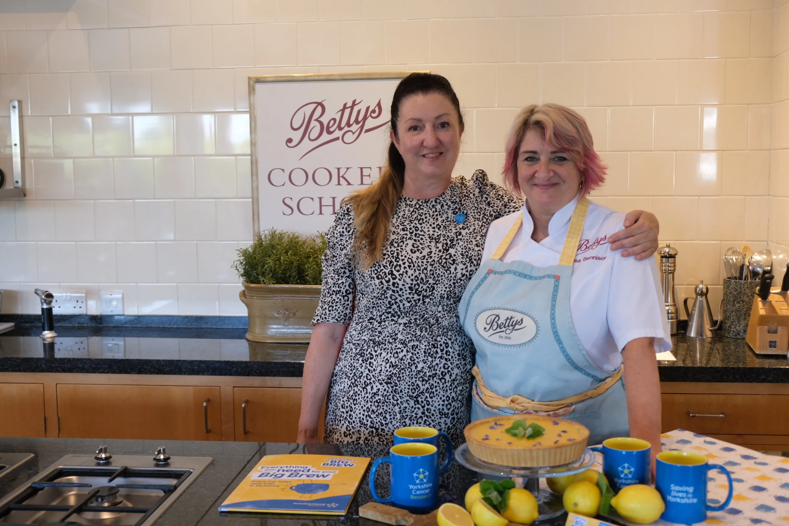 Ruth and Lisa from Bettys stand in Bettys Cookery School, with their arms around each other smiling at the camera. A lemon tart and Yorkshire Cancer Research merchandise is visible on the worktop in front.