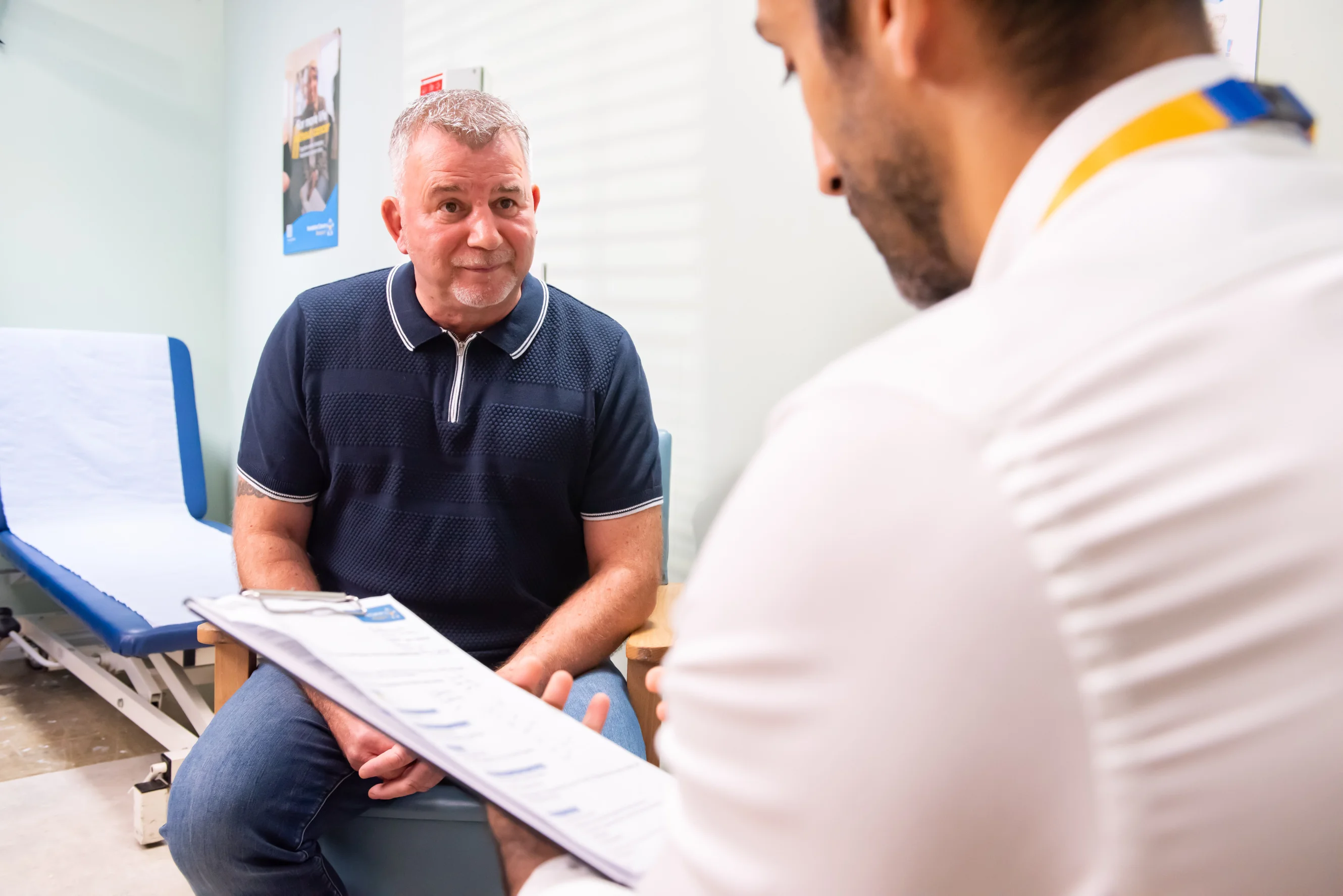 A patient sits in a medical setting while in conversation with a healthcare professional, who is wearing a Yorkshire Cancer Research lanyard and holding a clipboard.