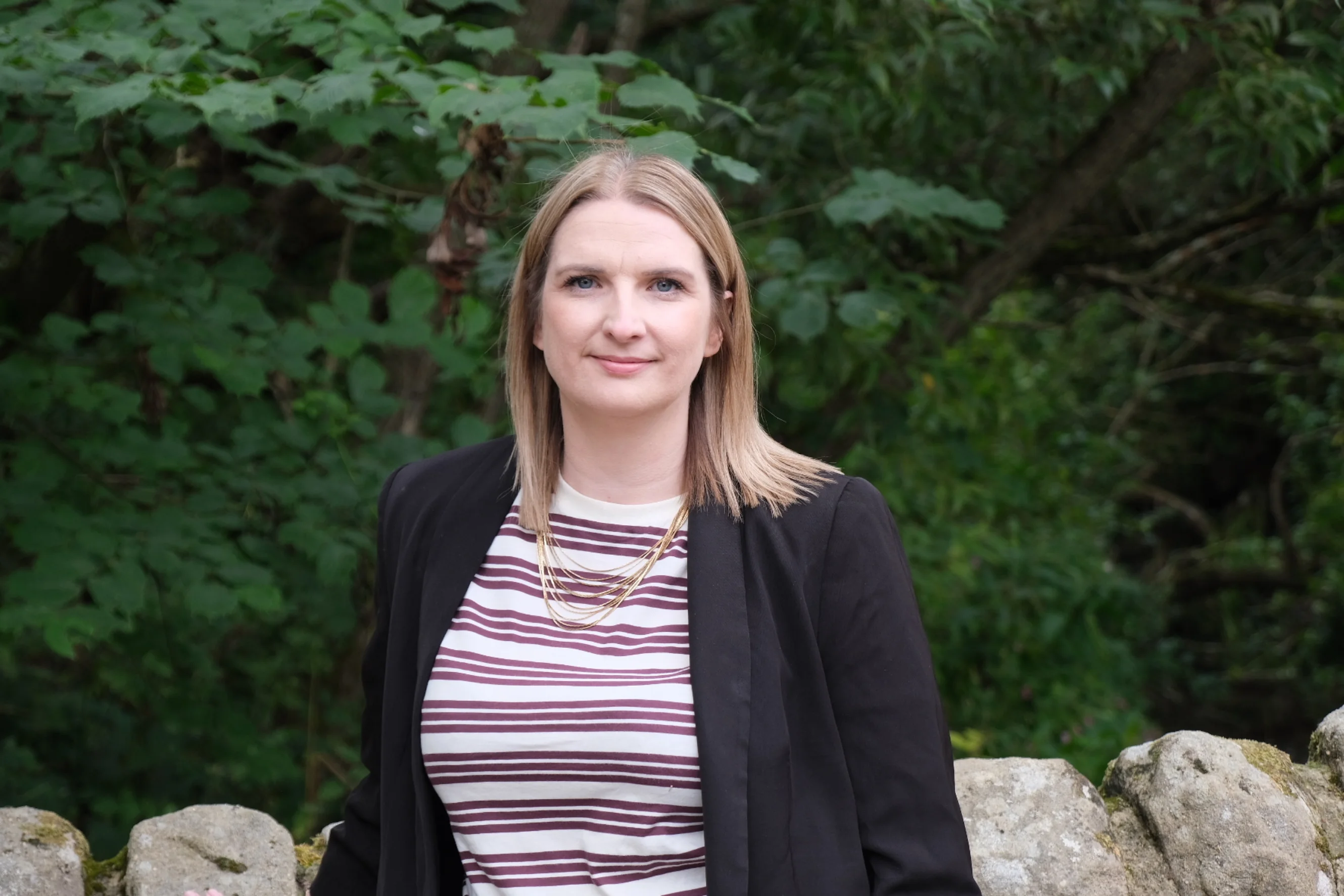 Beth Elias standing, facing the camera in a striped shirt, black blazer against a wall and trees